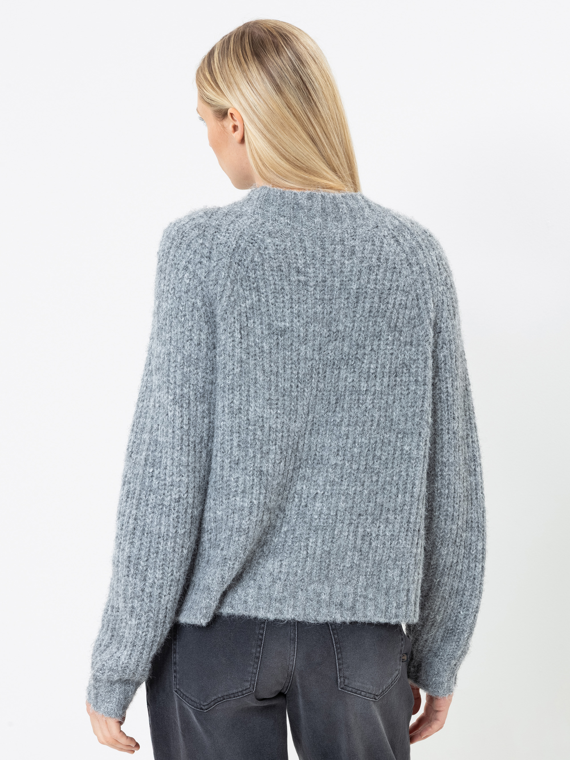 Eine Person mit glattem blondem Haar, von hinten gesehen, trägt einen strukturierten hellgrauen Strickpullover und dunkelgraue Jeans und steht vor einem schlichten weißen Hintergrund.