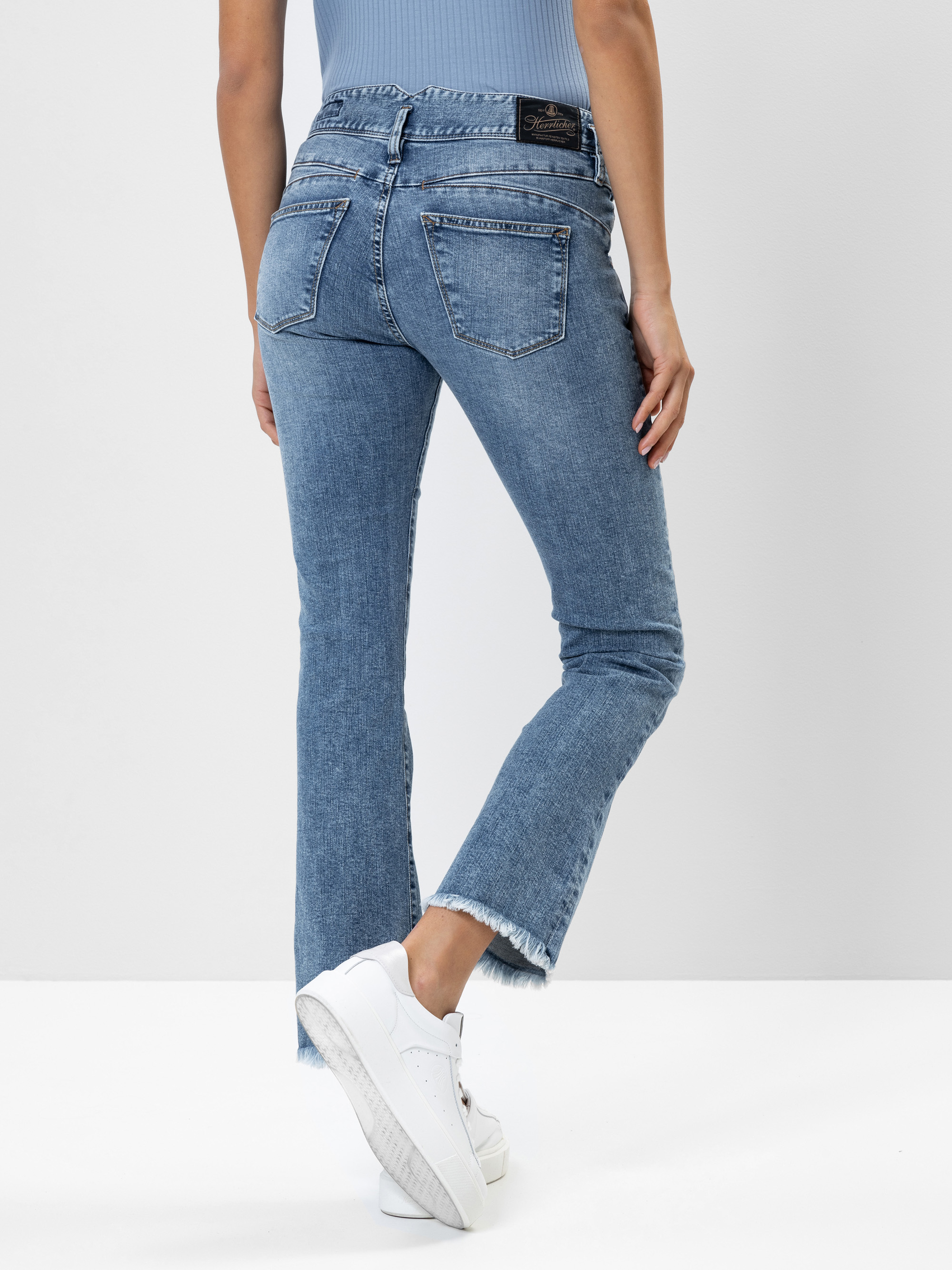 Eine Person, die eine hellblaue Jeans mit ausgefransten Säumen und ein blaues, geripptes Oberteil trägt, steht auf einem weißen Fußboden und wendet sich ab. Sie trägt weiße Turnschuhe.