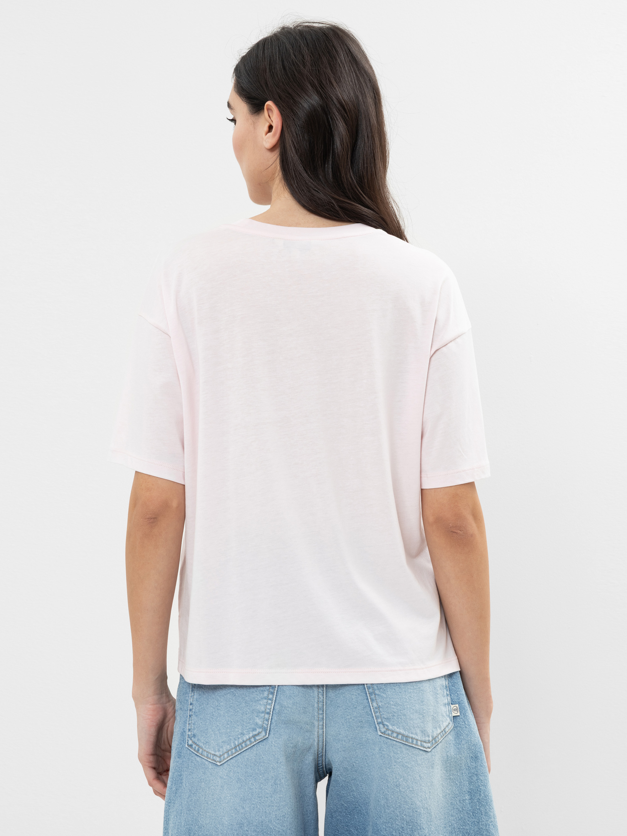 Eine Frau mit langen dunklen Haaren steht mit dem Rücken zur Kamera, trägt ein hellrosa kurzärmeliges T-Shirt und hellblaue Jeans vor einem schlichten weißen Hintergrund.