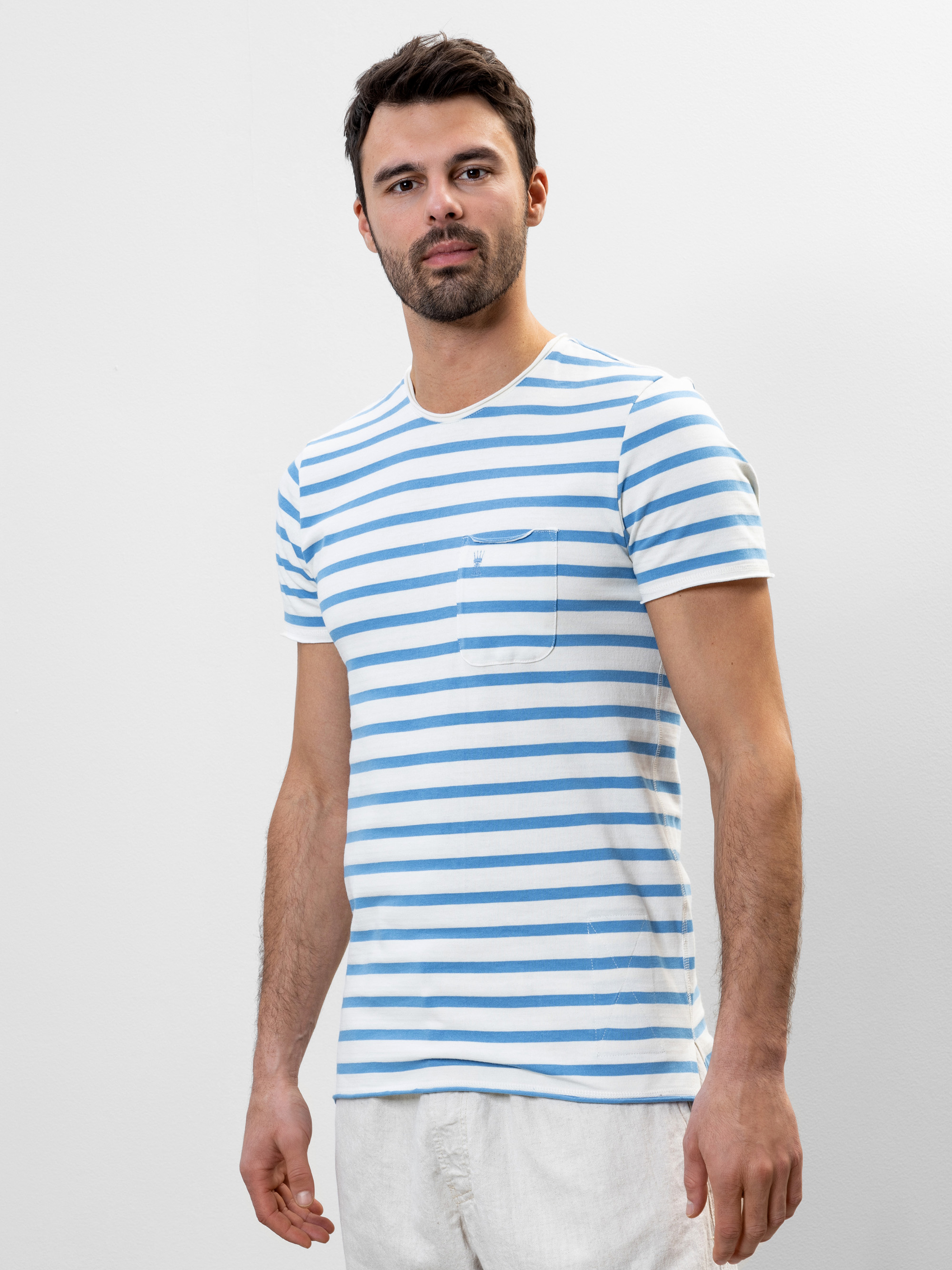 Ein Mann, der ein hellblau-weiß gestreiftes T-Shirt mit einer kleinen Brusttasche und eine weiße Hose trägt, steht vor einem schlichten hellen Hintergrund und schaut leicht zur Seite.