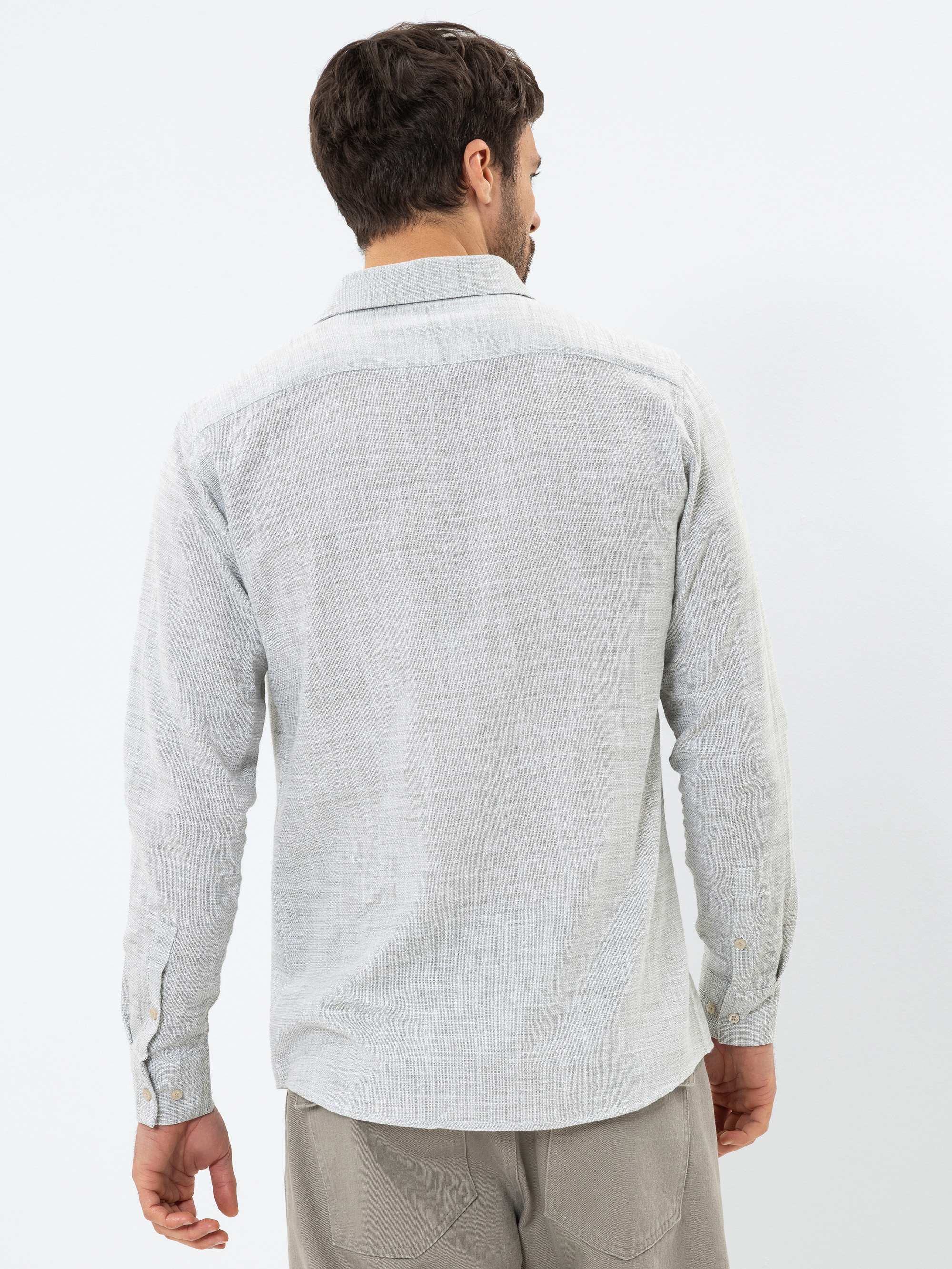 Ein Mann steht von der Kamera abgewandt in einem hellgrauen, langärmeligen Button-up-Hemd und einer hellgrauen Hose vor einem schlichten weißen Hintergrund.