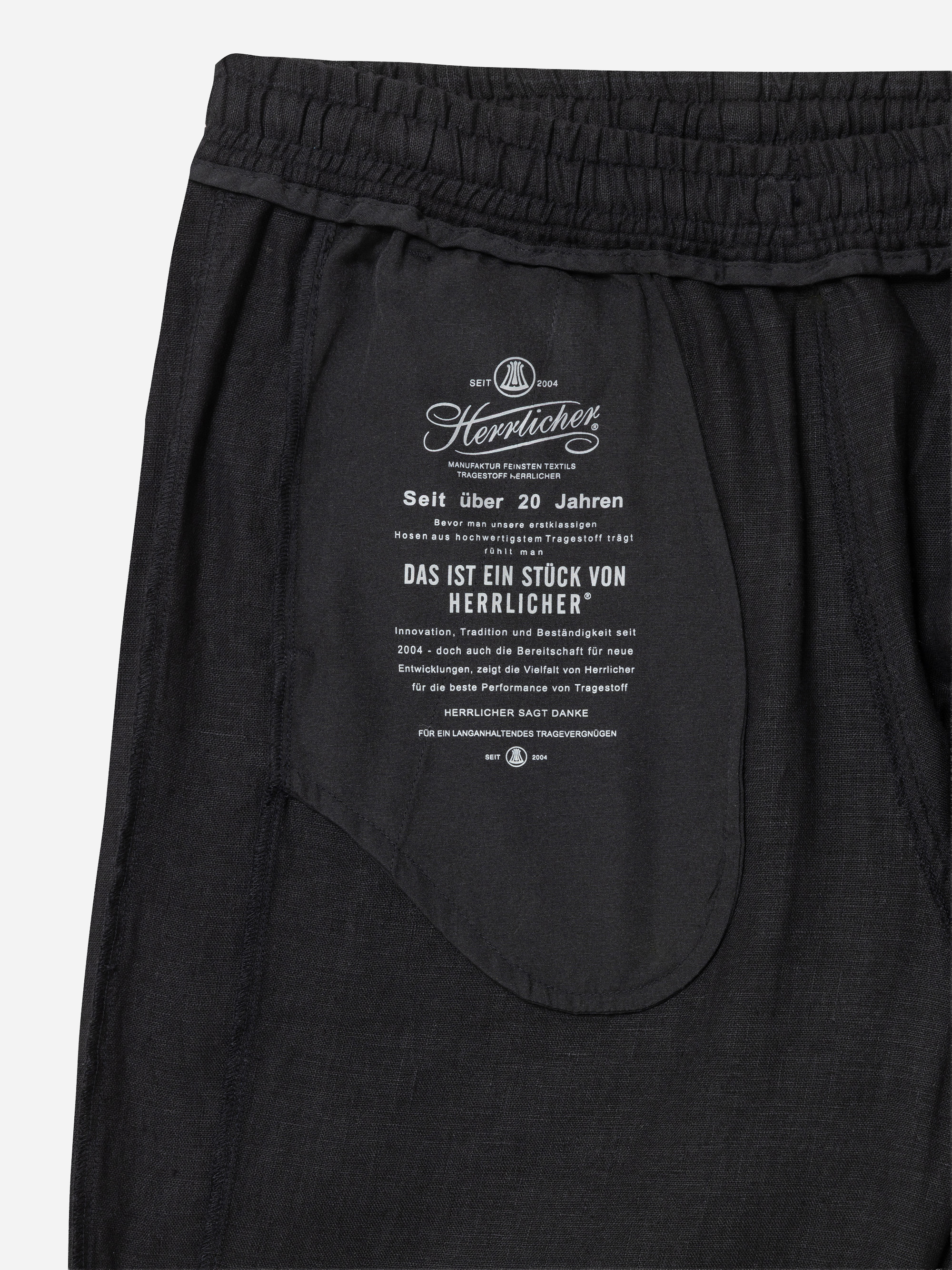 Nahaufnahme der schwarzen Hose mit der Innentasche, auf der ein weißer Text in deutscher Sprache über die Marke Herrlicher und ihre über 20-jährige Geschichte gedruckt ist. Der Bund ist elastisch und gerafft.