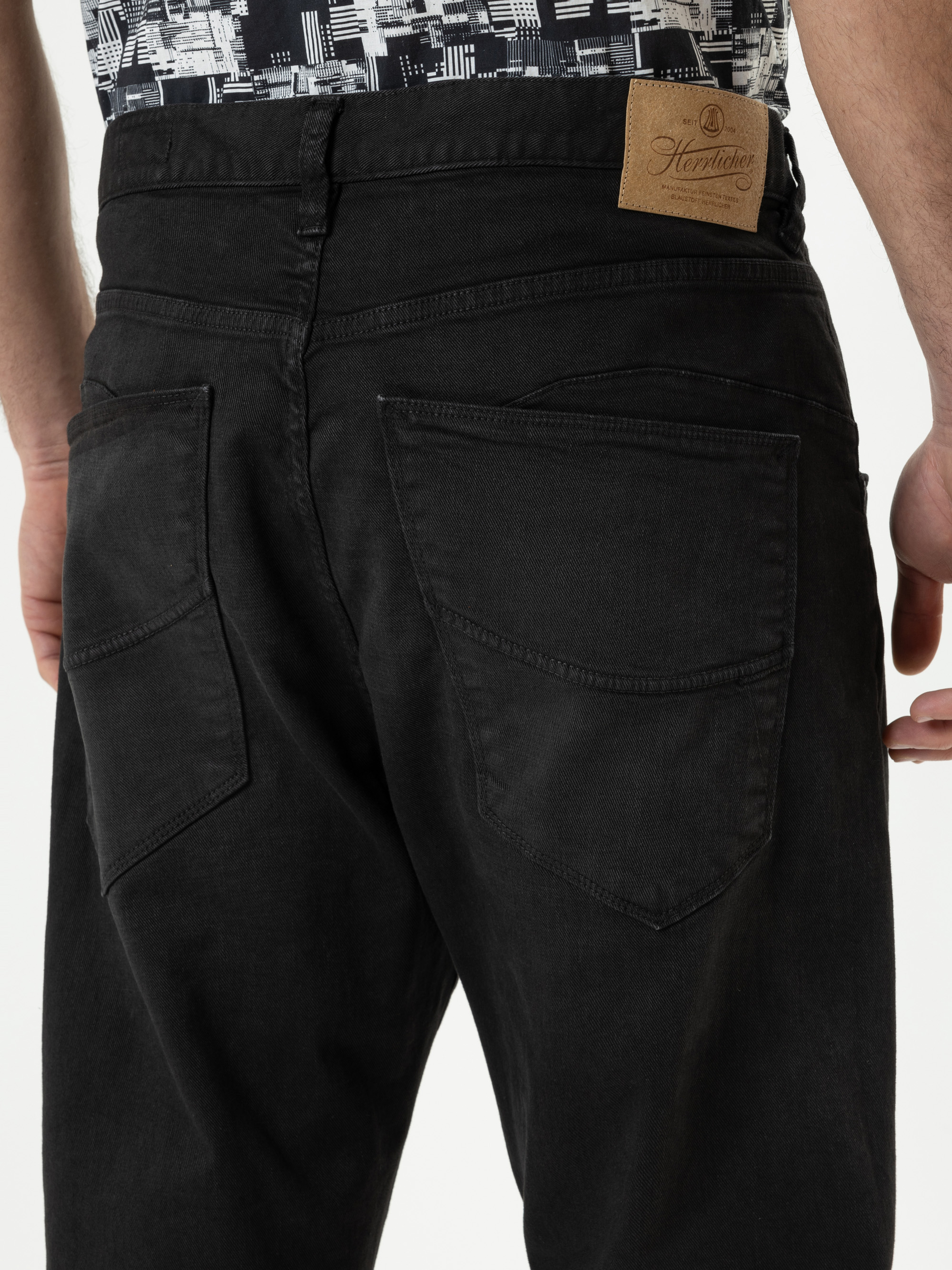 Nahaufnahme einer Person, die eine schwarze Jeans trägt, wobei die Gesäßtaschen und ein hellbrauner Markenaufnäher am Bund zu sehen sind. Die Person steht mit entspannten Armen an der Seite.