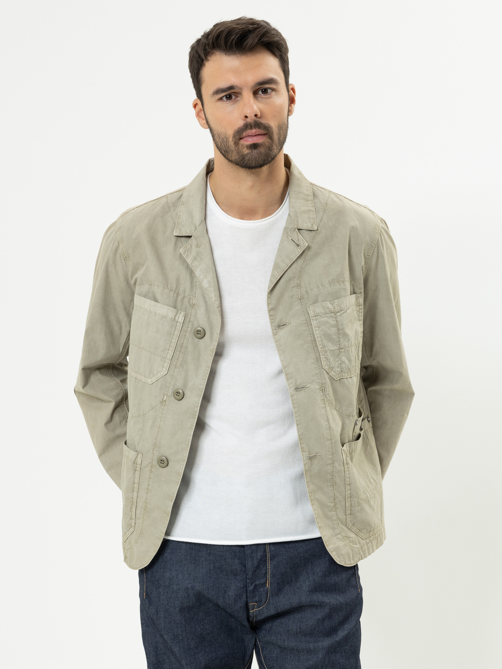 Ein Mann mit kurzen dunklen Haaren und einem Bart trägt eine helle khakifarbene Jacke über einem weißen T-Shirt und dunkle Jeans. Er steht mit den Händen auf dem Rücken vor einem schlichten weißen Hintergrund.