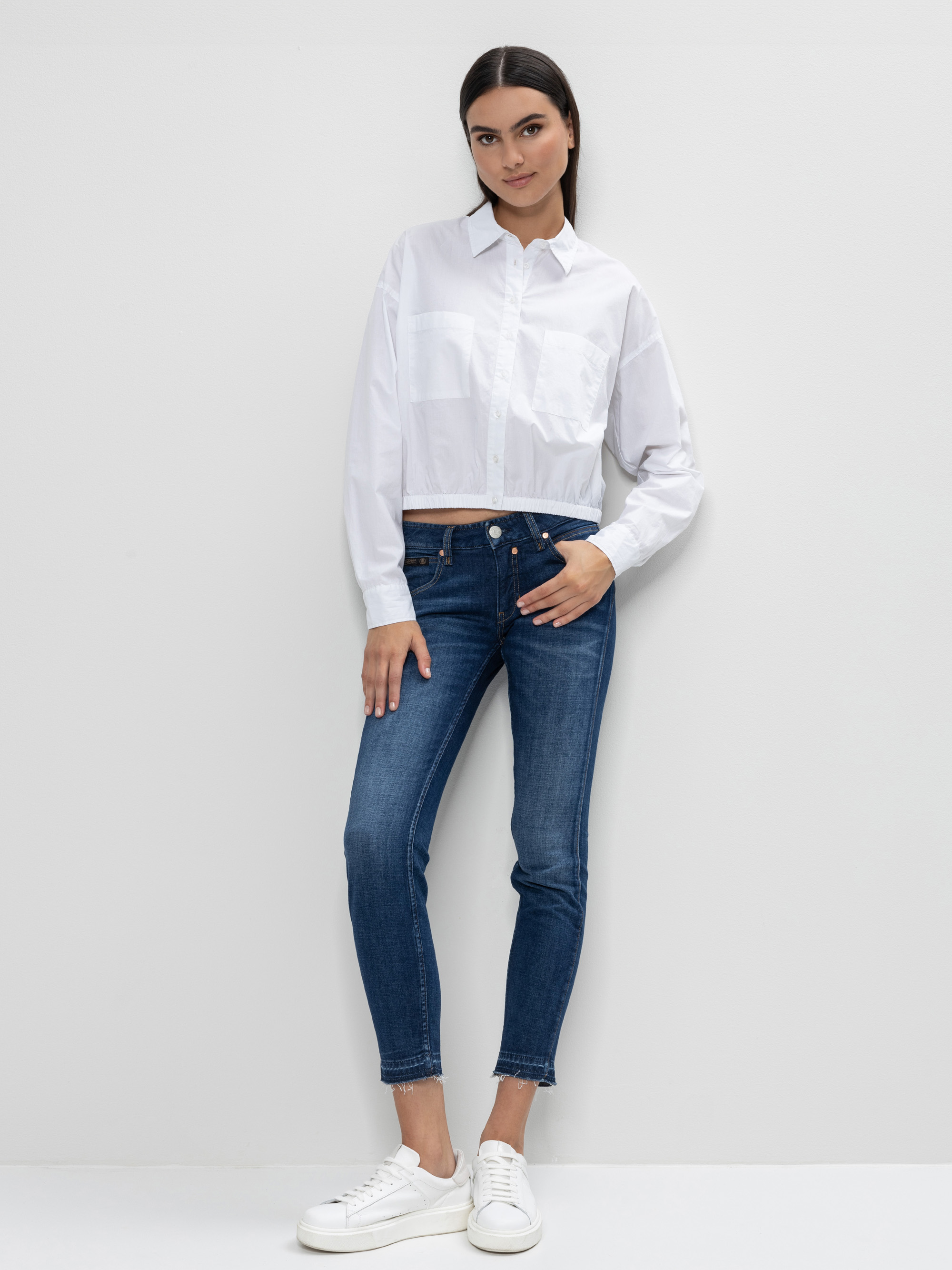 Eine Frau steht vor einer weißen Wand und trägt ein weißes, geknöpftes Hemd, blaue Skinny Jeans und weiße Turnschuhe. Sie hat glattes dunkles Haar und posiert mit einer Hand in der Tasche und einem angewinkelten Bein.