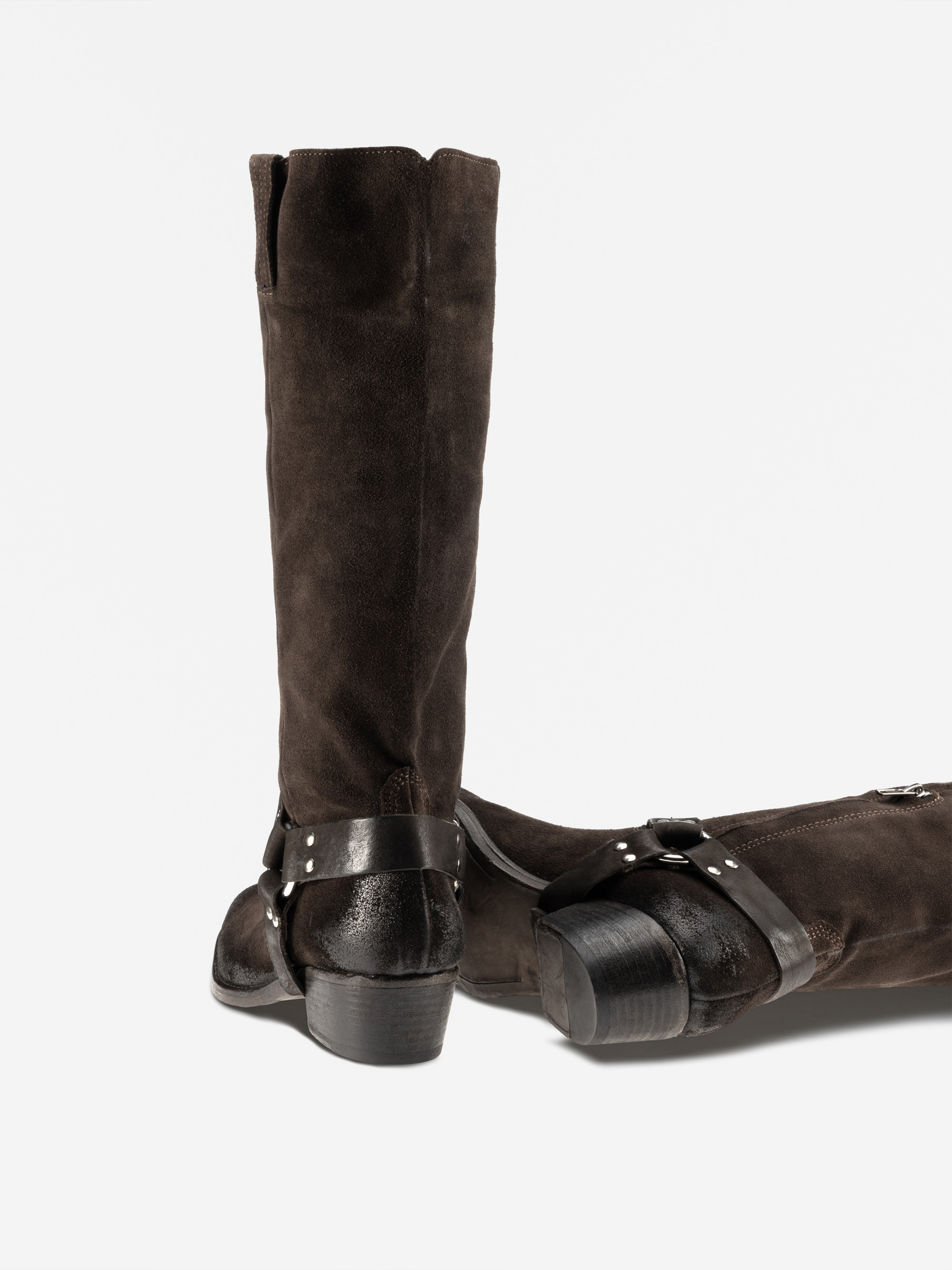 Ein Paar hohe, dunkelbraune Wildlederstiefel mit schwarzen Lederriemen und silbernen Nieten, von denen einer aufrecht steht und der andere auf der Seite liegt, vor einem schlichten weißen Hintergrund.