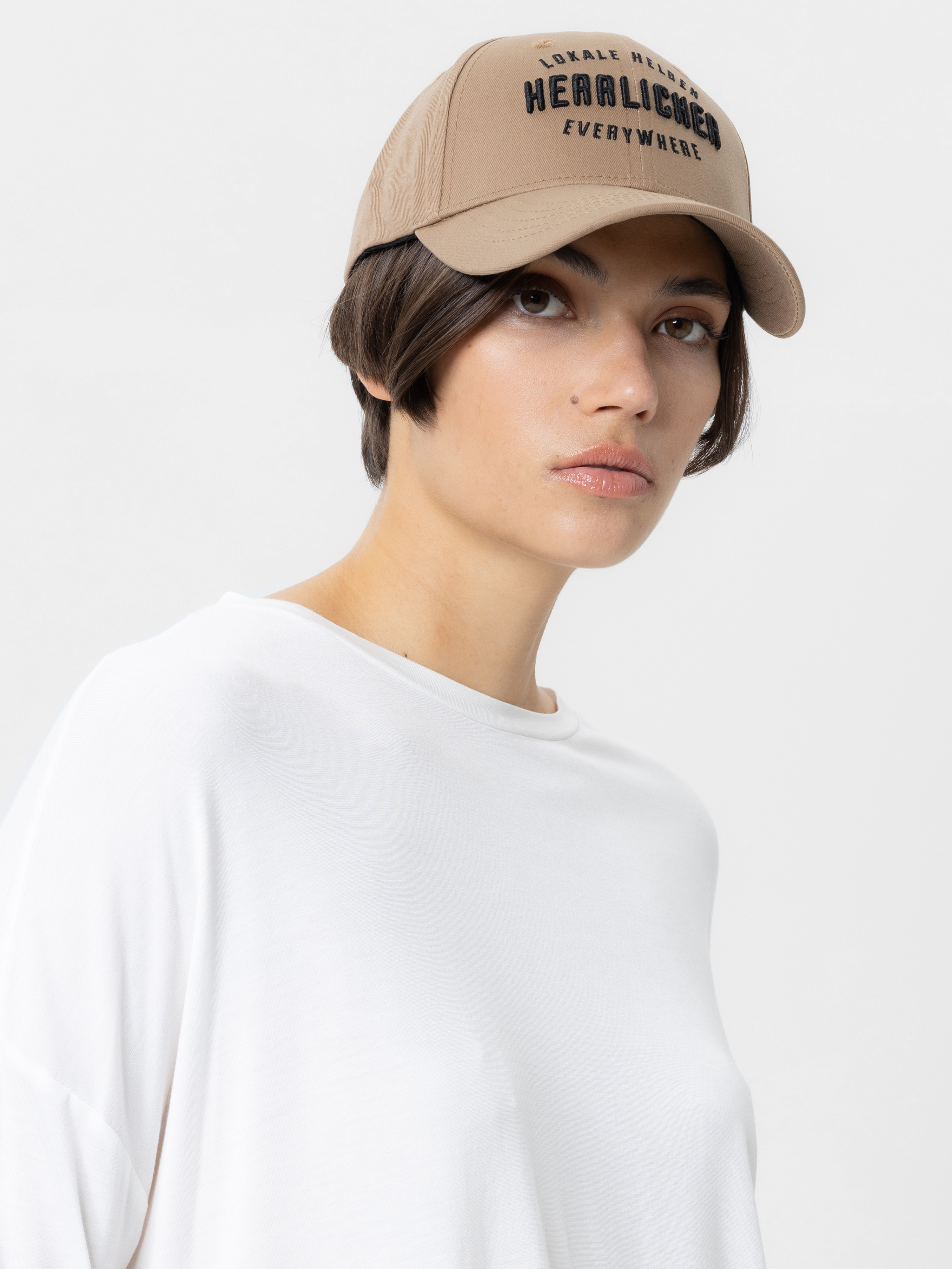 Eine Frau mit kurzen dunklen Haaren trägt eine hellbraune Baseballkappe und ein lockeres weißes Oberteil und blickt mit neutralem Gesichtsausdruck vor einem schlichten hellen Hintergrund leicht an der Kamera vorbei.