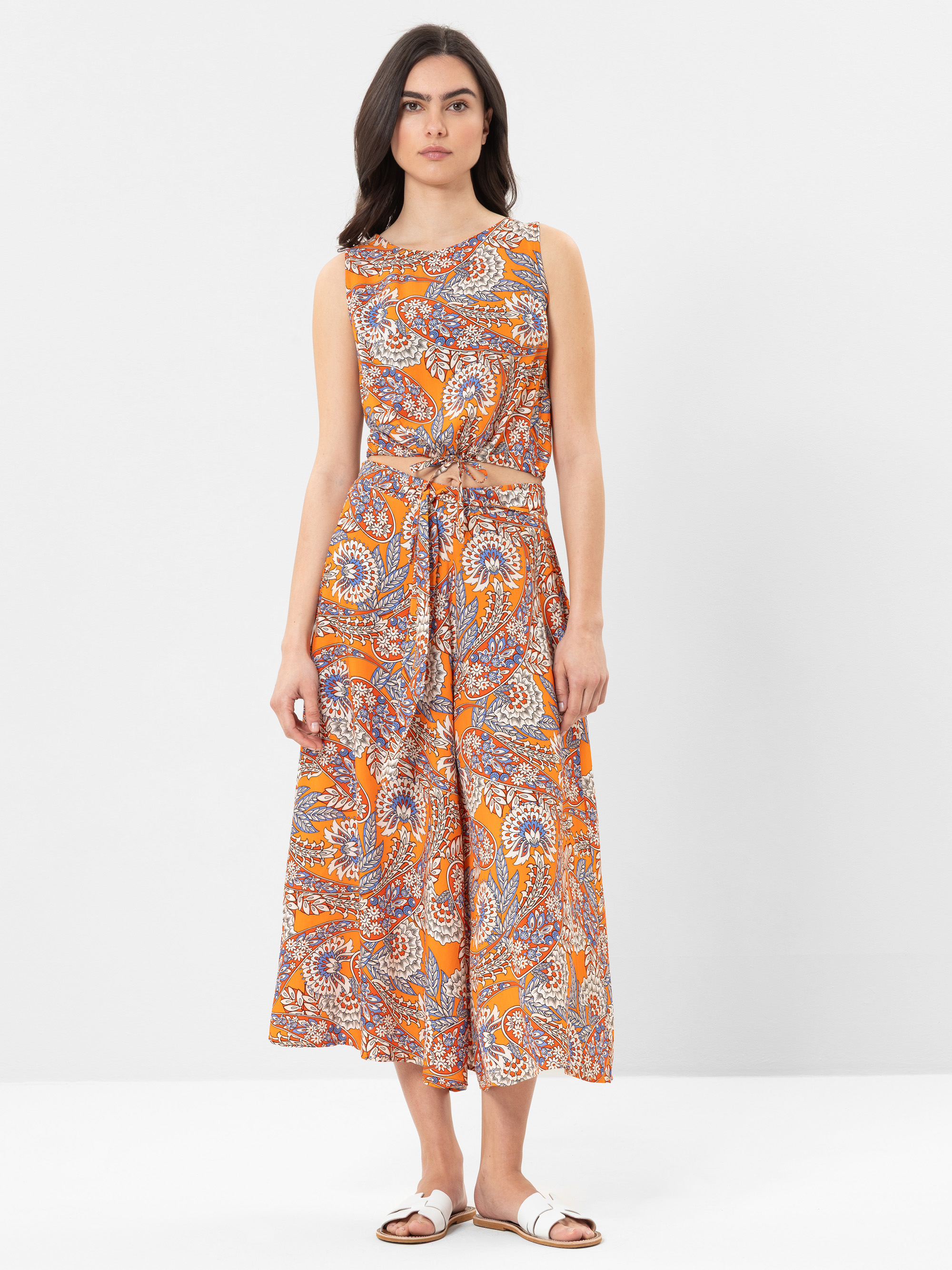 Eine Frau steht vor einem schlichten weißen Hintergrund und trägt ein ärmelloses, knöchellanges orangefarbenes Kleid mit einem bunten Paisleymuster und einem passenden Gürtel. Sie hat langes dunkles Haar und trägt weiße Sandalen.