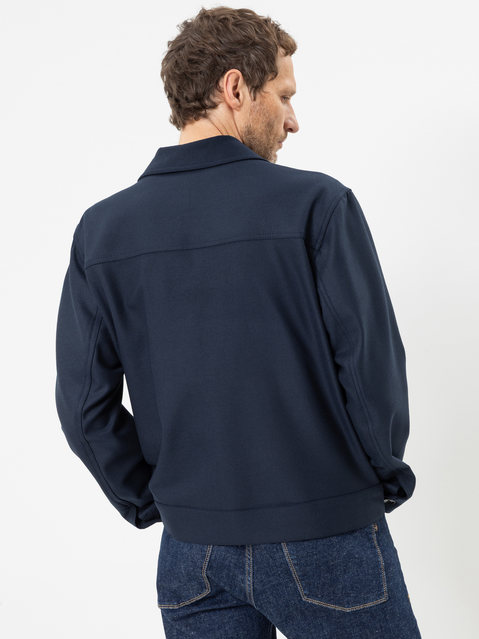 Ein Mann in einer dunkelblauen Jacke und blauen Jeans steht mit dem Rücken zur Kamera und blickt vor einem schlichten weißen Hintergrund nach links.