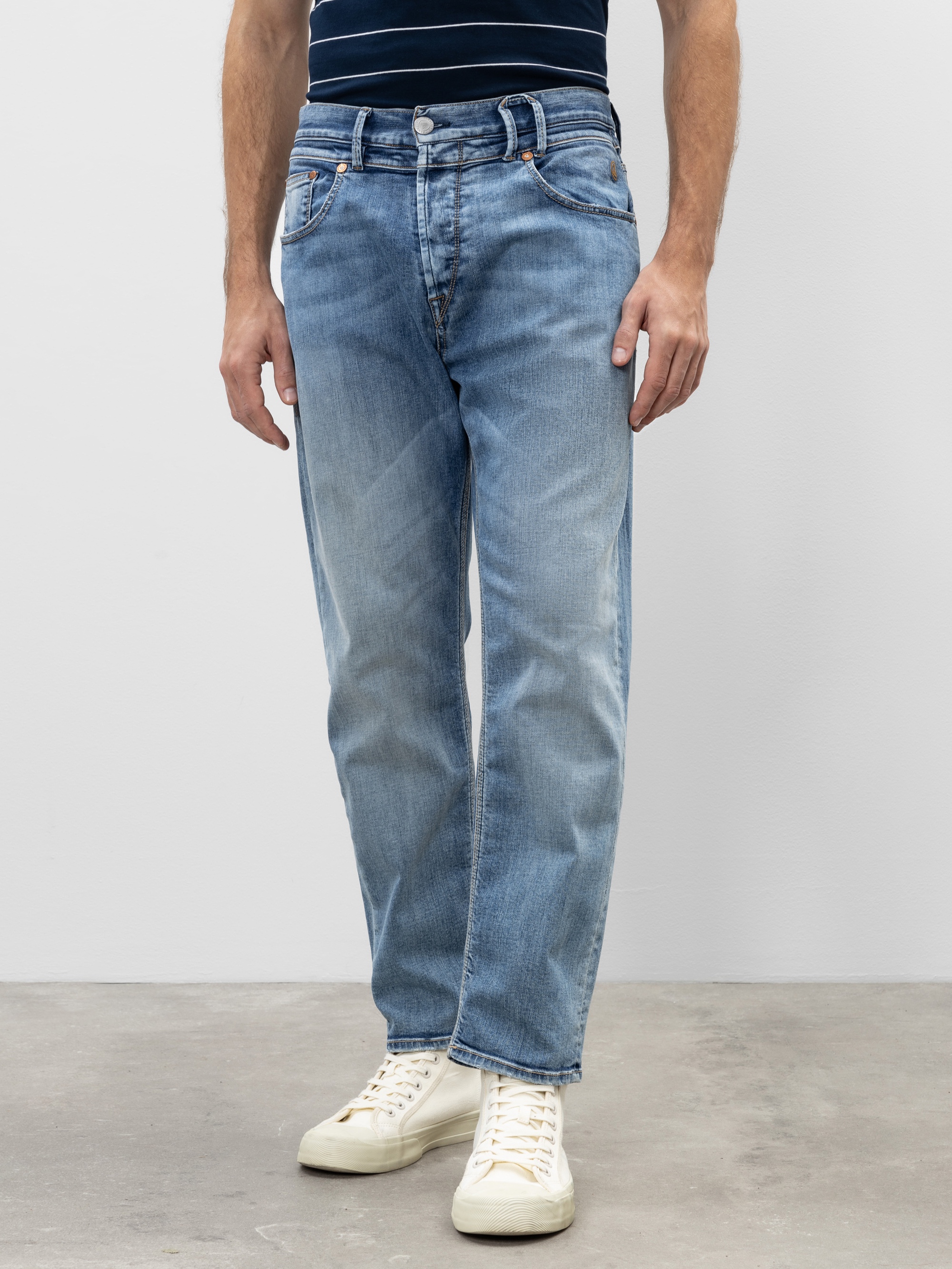 Eine Person in hellblauen Jeans mit geradem Bein und weißen Turnschuhen, die auf einem grauen Boden vor einer schlichten weißen Wand steht. Nur die untere Hälfte des Körpers ist sichtbar.