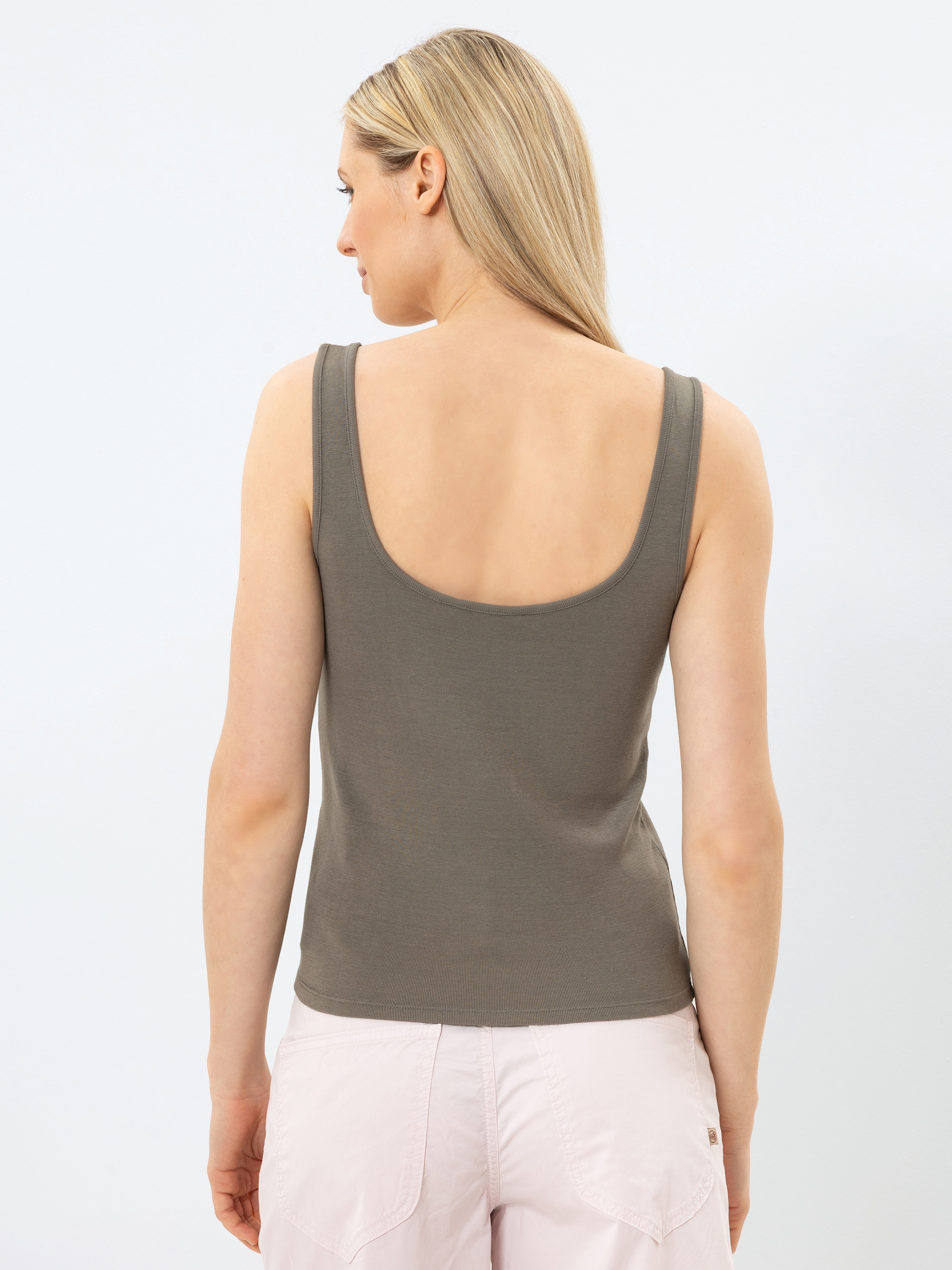 Eine Frau mit glattem, blondem Haar steht von der Kamera abgewandt, trägt ein taupefarbenes Tank-Top und hellrosa Shorts vor einem schlichten, hellen Hintergrund.