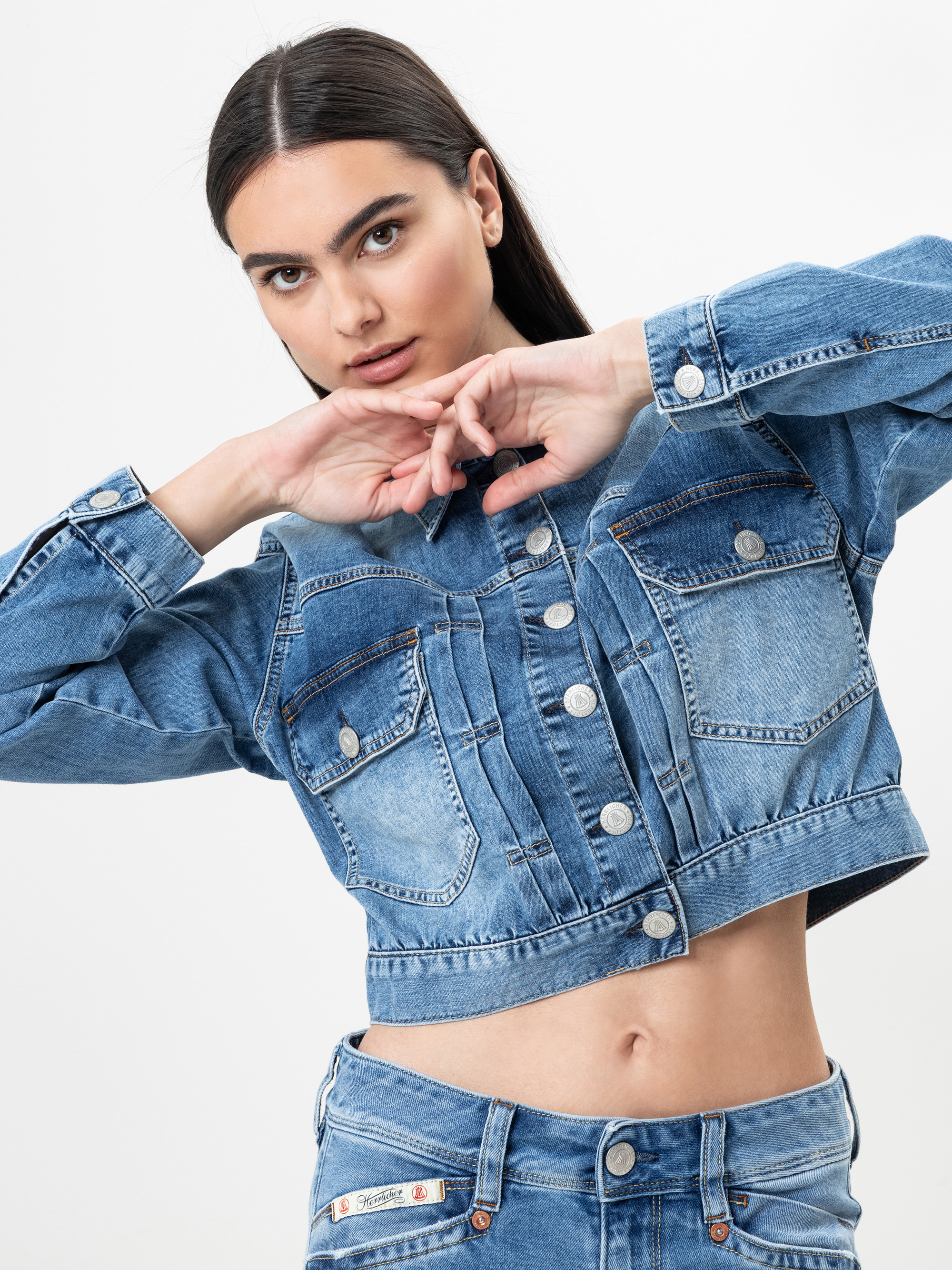 Eine Frau mit langen dunklen Haaren posiert in einer abgeschnittenen Jeansjacke und Jeans vor einem schlichten weißen Hintergrund, hebt ihre Arme und blickt selbstbewusst in die Kamera.