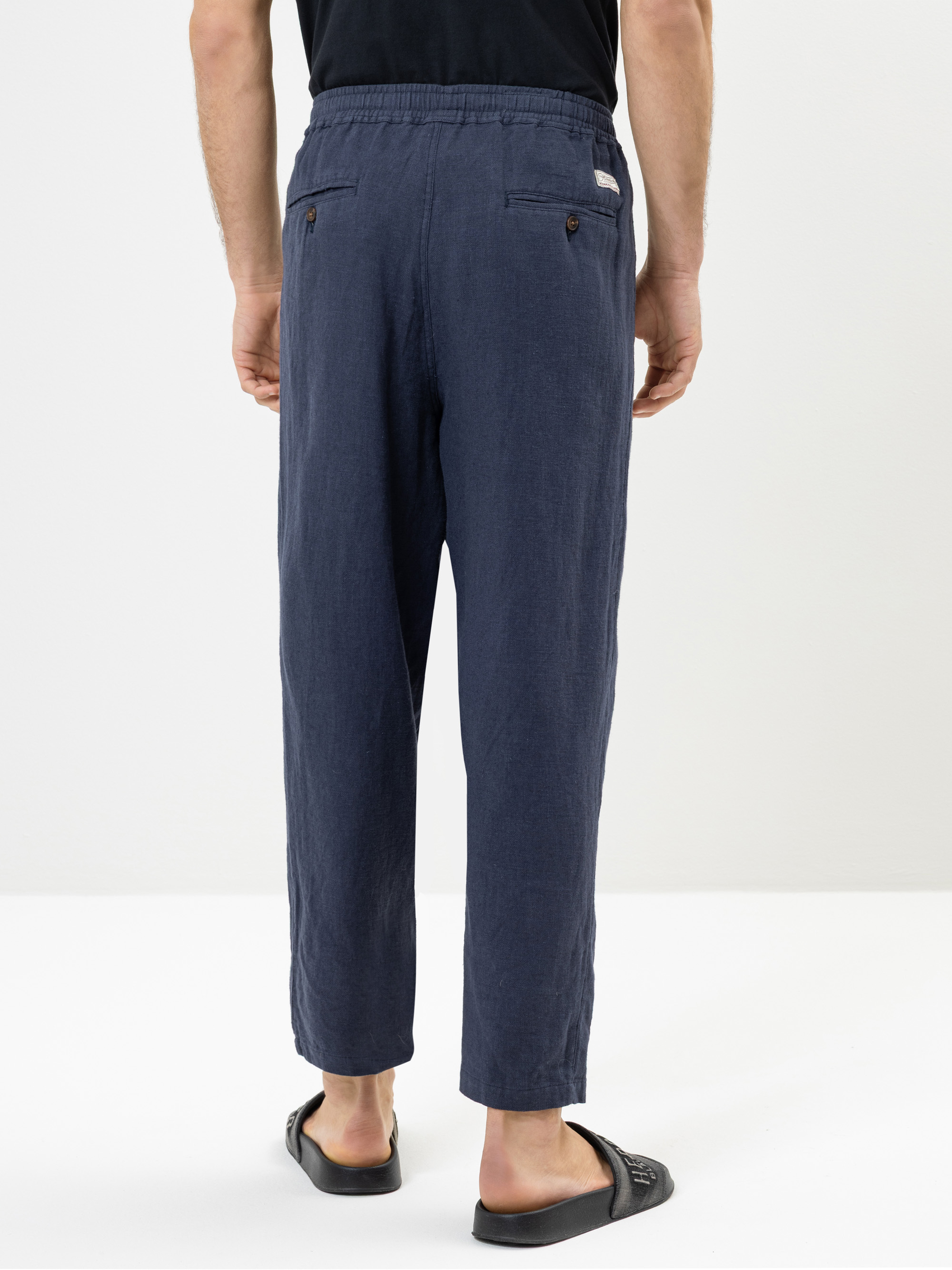 Eine Person steht mit dem Gesicht nach hinten und trägt eine marineblaue, locker sitzende Hose mit elastischem Bund und schwarze Sandalen. Die Hose hat Gesäßtaschen mit Knöpfen, und die Person trägt ein schwarzes Hemd. Der Hintergrund ist einfarbig w