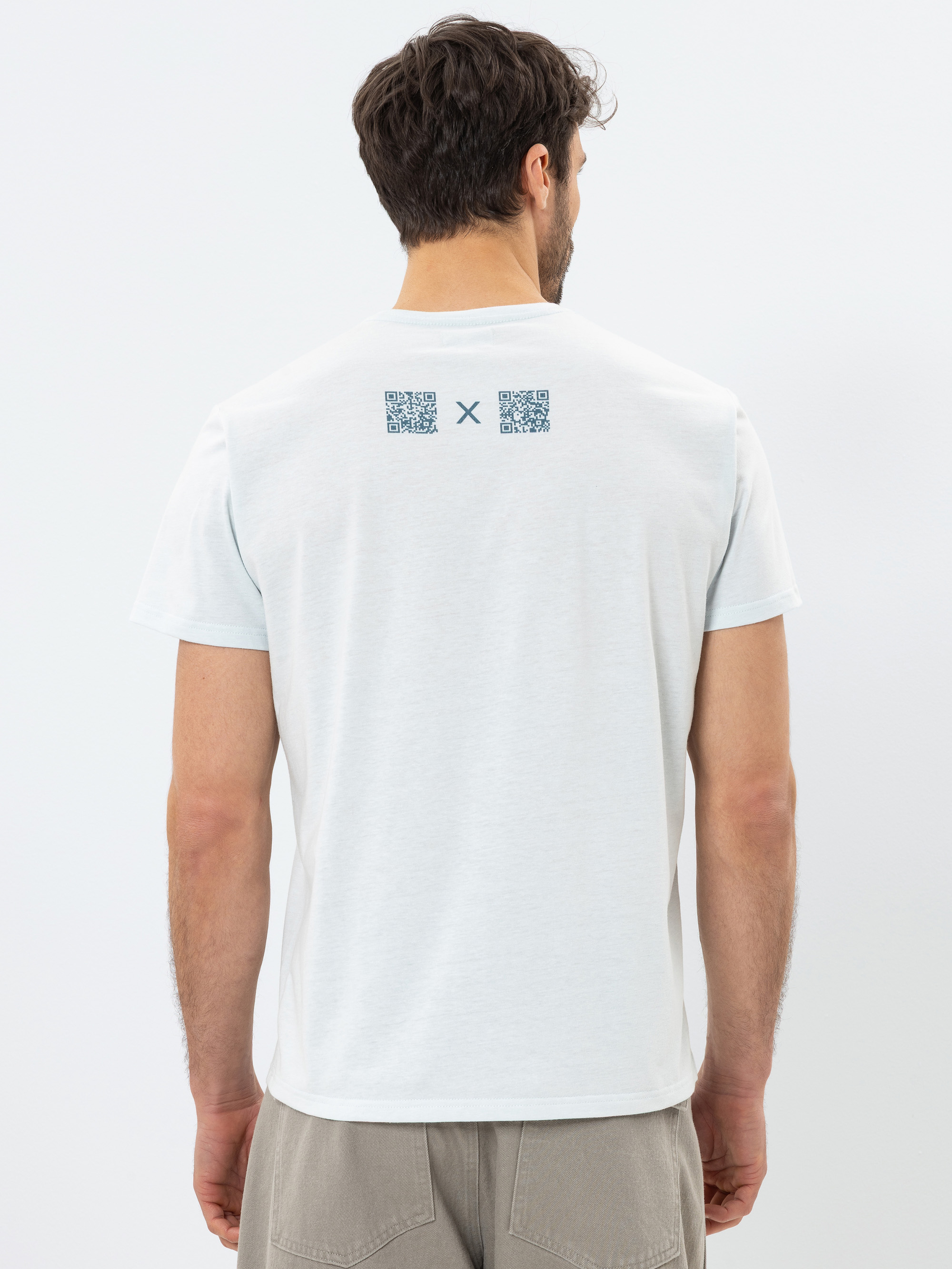 Ein Mann, der ein helles T-Shirt und beigefarbene Shorts trägt, ist von hinten zu sehen. Auf dem T-Shirt sind auf dem oberen Rücken zwei QR-Codes und dazwischen ein kleines X aufgedruckt. Der Hintergrund ist einfarbig weiß.