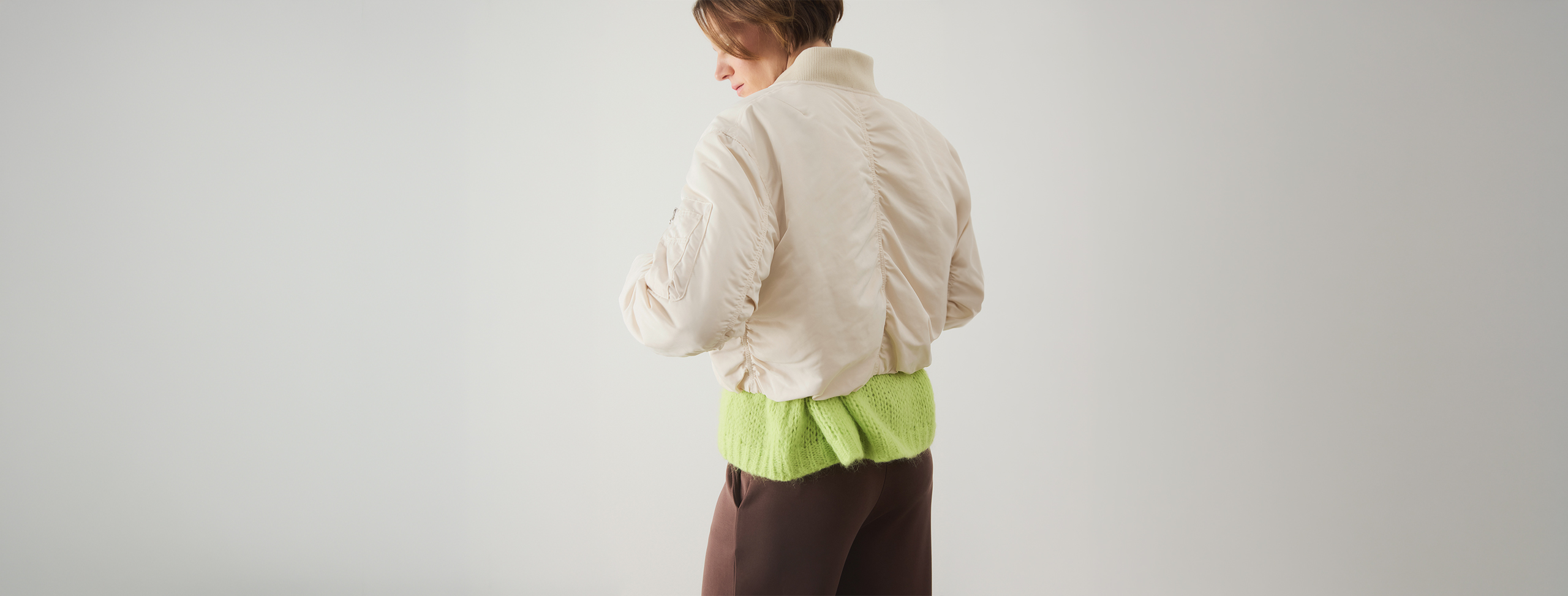 Eine Person steht mit dem Gesicht zur Wand und trägt eine hellbeige Jacke, einen hellgrünen strukturierten Pullover und eine braune Hose vor einem schlichten hellen Hintergrund.