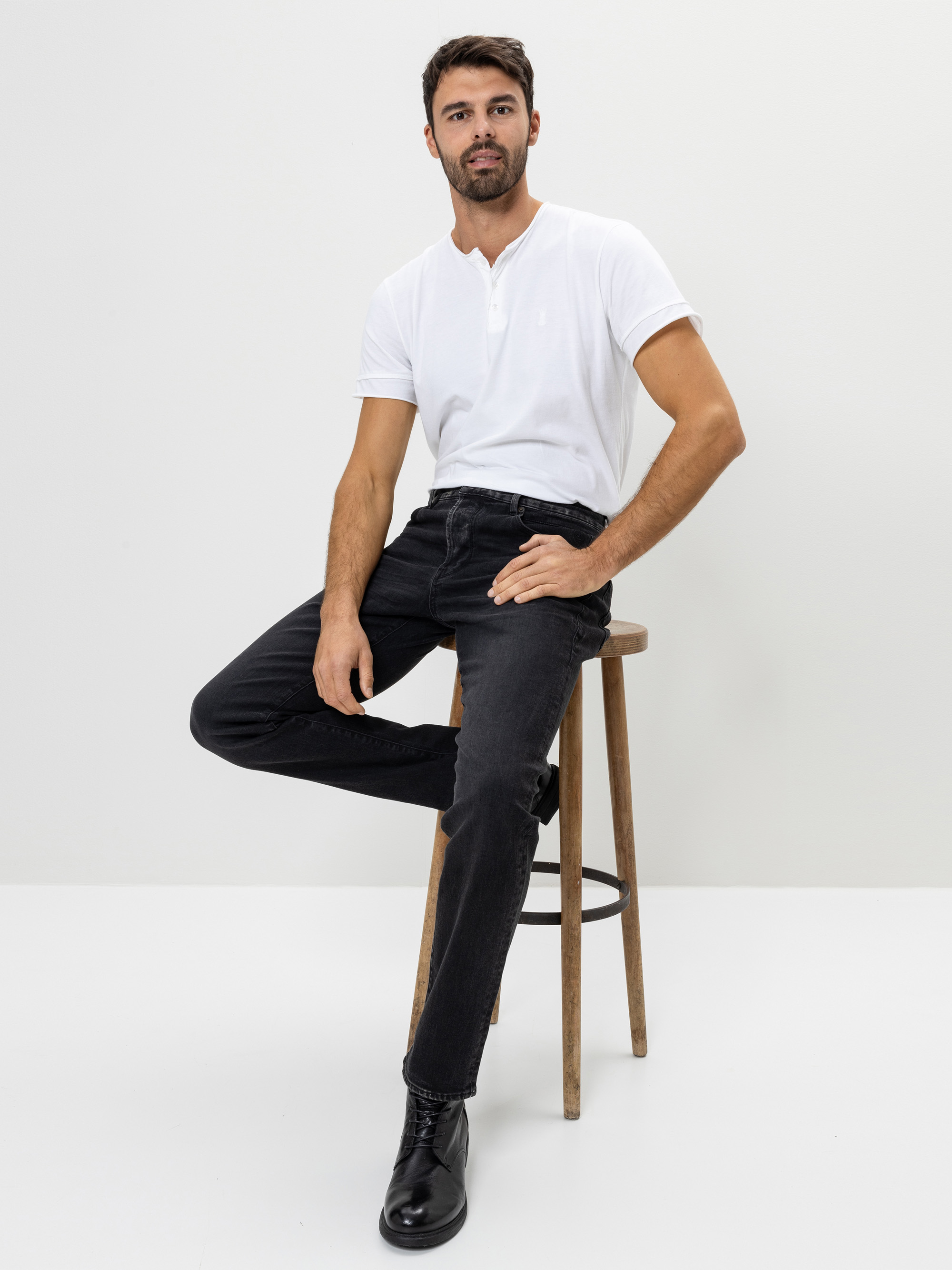 Ein Mann mit kurzen dunklen Haaren und Bart sitzt auf einem Holzschemel vor einem schlichten weißen Hintergrund. Er trägt ein weißes kurzärmeliges Hemd, schwarze Jeans und schwarze Schuhe. Ein Fuß steht auf dem Boden, der andere stützt sich auf ein