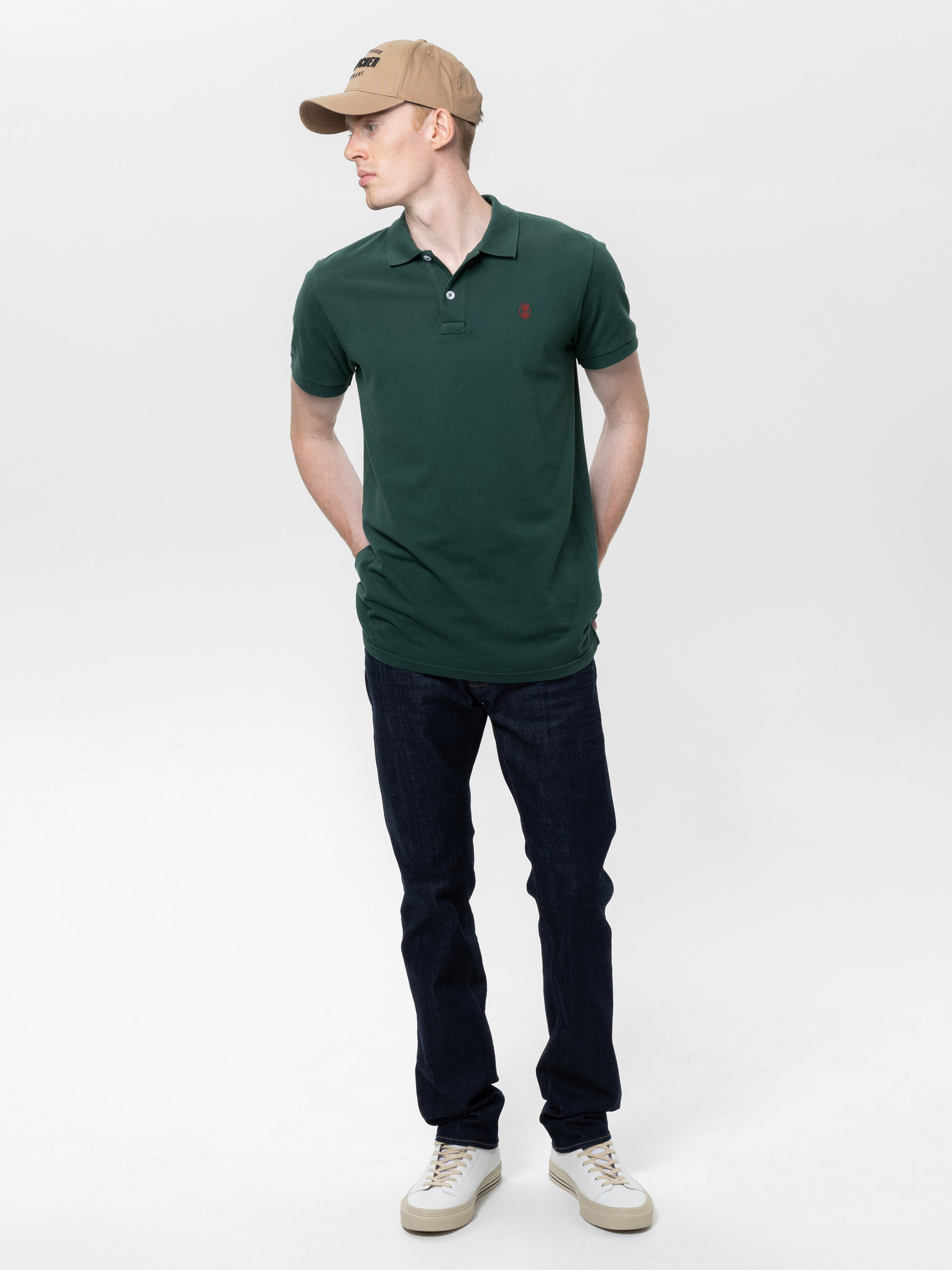 Eine Person, die ein grünes Polohemd, dunkle Jeans, weiße Turnschuhe und eine beigefarbene Kappe trägt, steht mit den Händen auf dem Rücken und blickt zur Seite vor einem schlichten weißen Hintergrund.