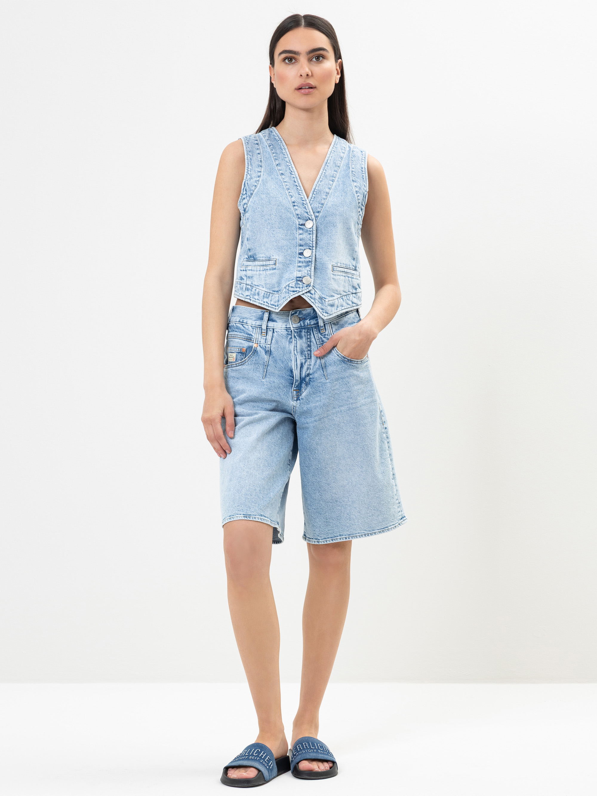 Eine Frau steht vor einem schlichten Hintergrund und trägt eine ärmellose, hochgeknöpfte Jeansweste und knielange, lockere Jeansshorts. Sie hat langes, glattes Haar und trägt dunkle Slide-Sandalen. Eine Hand steckt in ihrer Tasche.