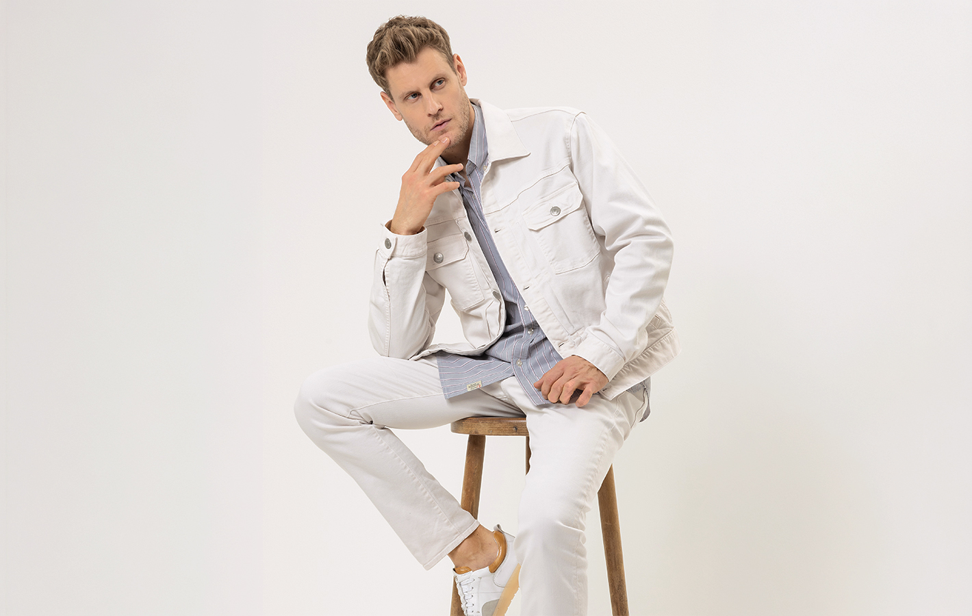 Ein Mann in einer hellen Jeansjacke und einer hellen Jeanshose sitzt auf einem Holzhocker vor einem weißen Hintergrund, stützt eine Hand am Kinn ab und blickt nachdenklich in die Kamera.