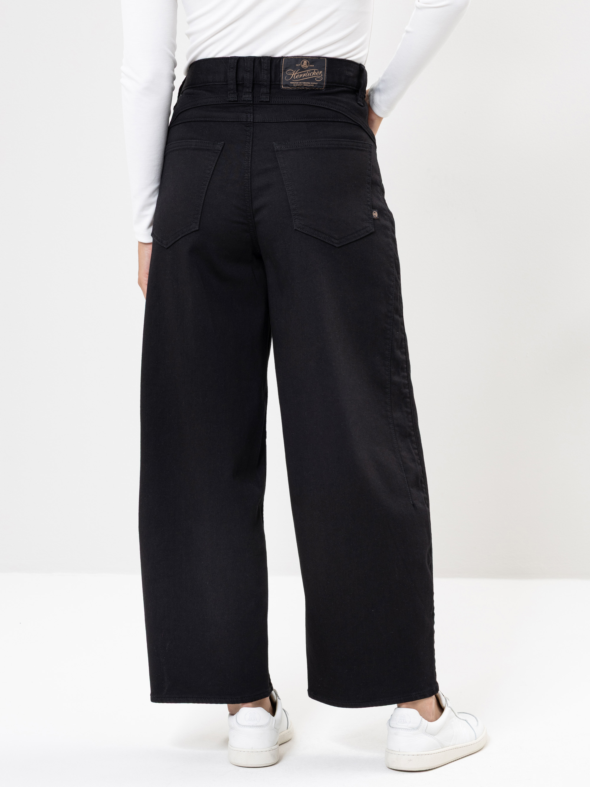 Eine Person, die eine schwarze Jeans mit hoher Taille und weitem Bein, ein weißes langärmeliges Oberteil und weiße Turnschuhe trägt, wird von hinten vor einem schlichten hellen Hintergrund gezeigt.