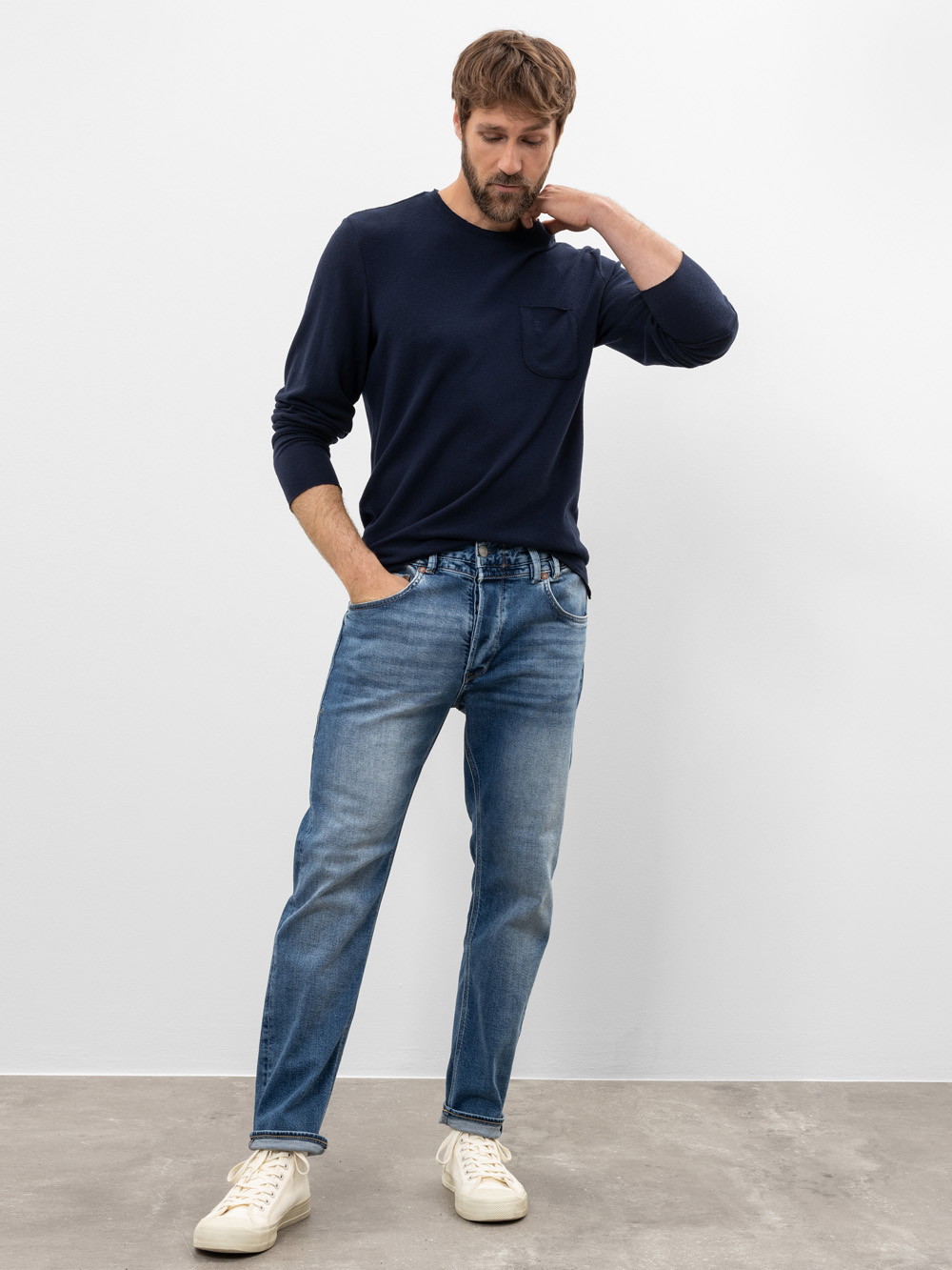 Ein Mann steht in einem Innenraum vor einer schlichten weißen Wand. Er trägt ein marineblaues langärmeliges Hemd, blaue Jeans und weiße Turnschuhe. Eine Hand steckt in seiner Hosentasche, während die andere seinen Nacken berührt, und er schaut nachd