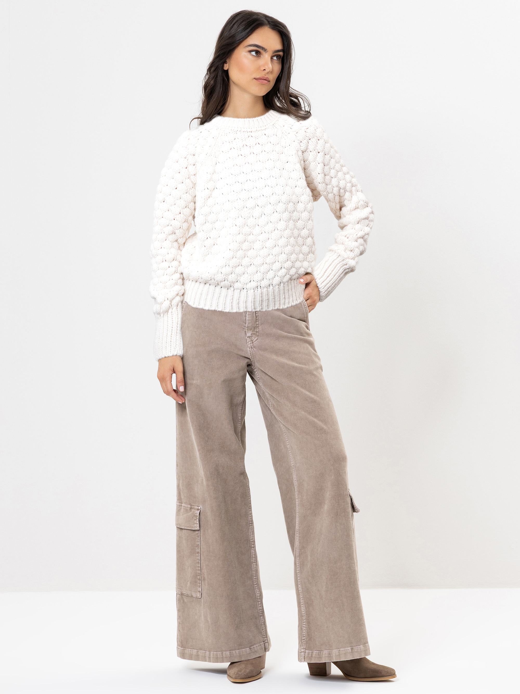 Eine Frau steht vor einem schlichten weißen Hintergrund und trägt einen strukturierten weißen Pullover, eine beigefarbene Cargohose mit weitem Bein und hellbraune Stiefel. Sie hat langes dunkles Haar und blickt mit einer Hand in der Tasche zur Seite.