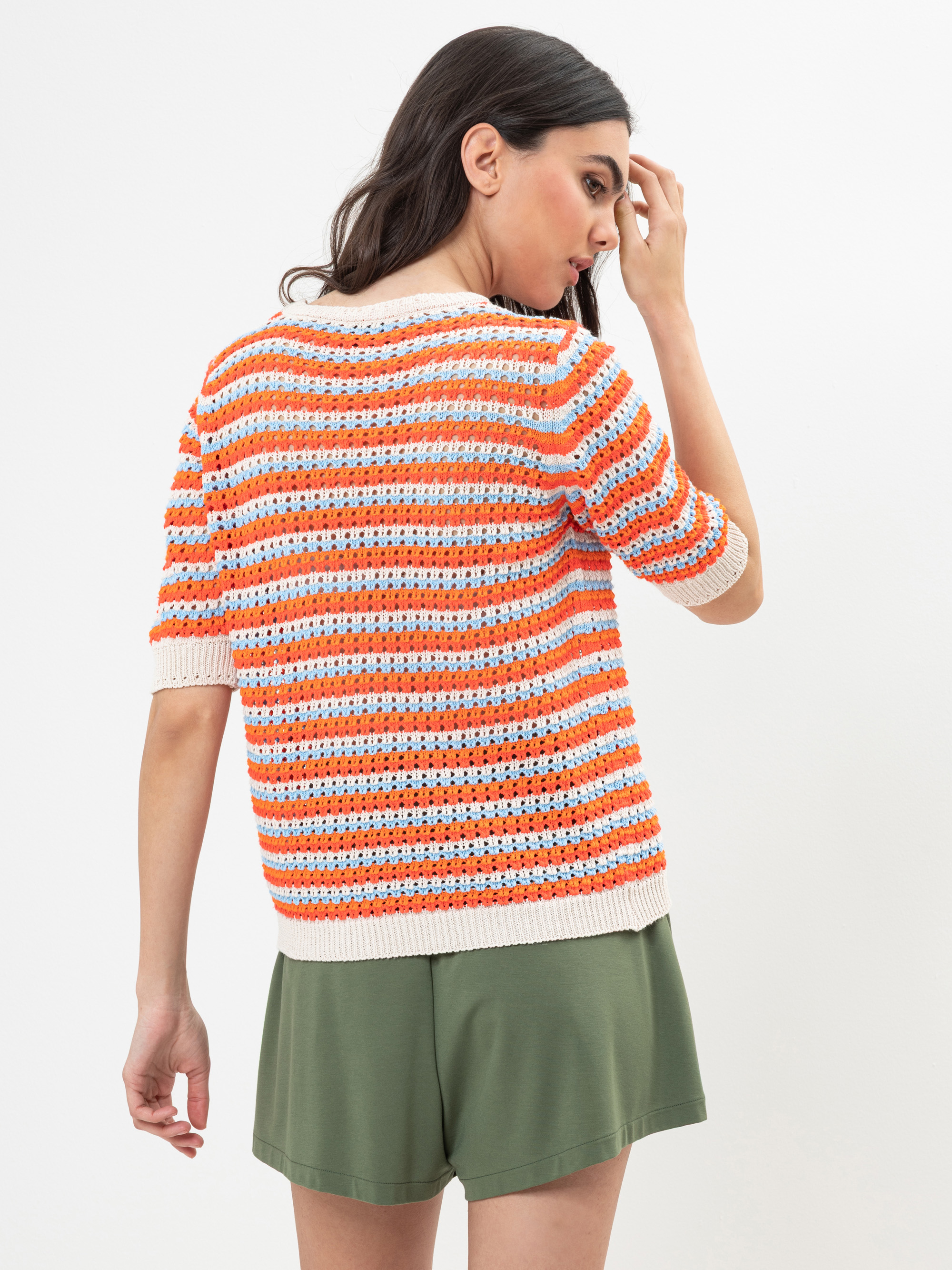 Eine Frau mit langen dunklen Haaren trägt einen orange, weiß und hellblau gestreiften, kurzärmeligen Pullover und olivgrüne Shorts. Sie steht vor einem schlichten weißen Hintergrund, schaut weg und berührt mit einer Hand ihr Gesicht.