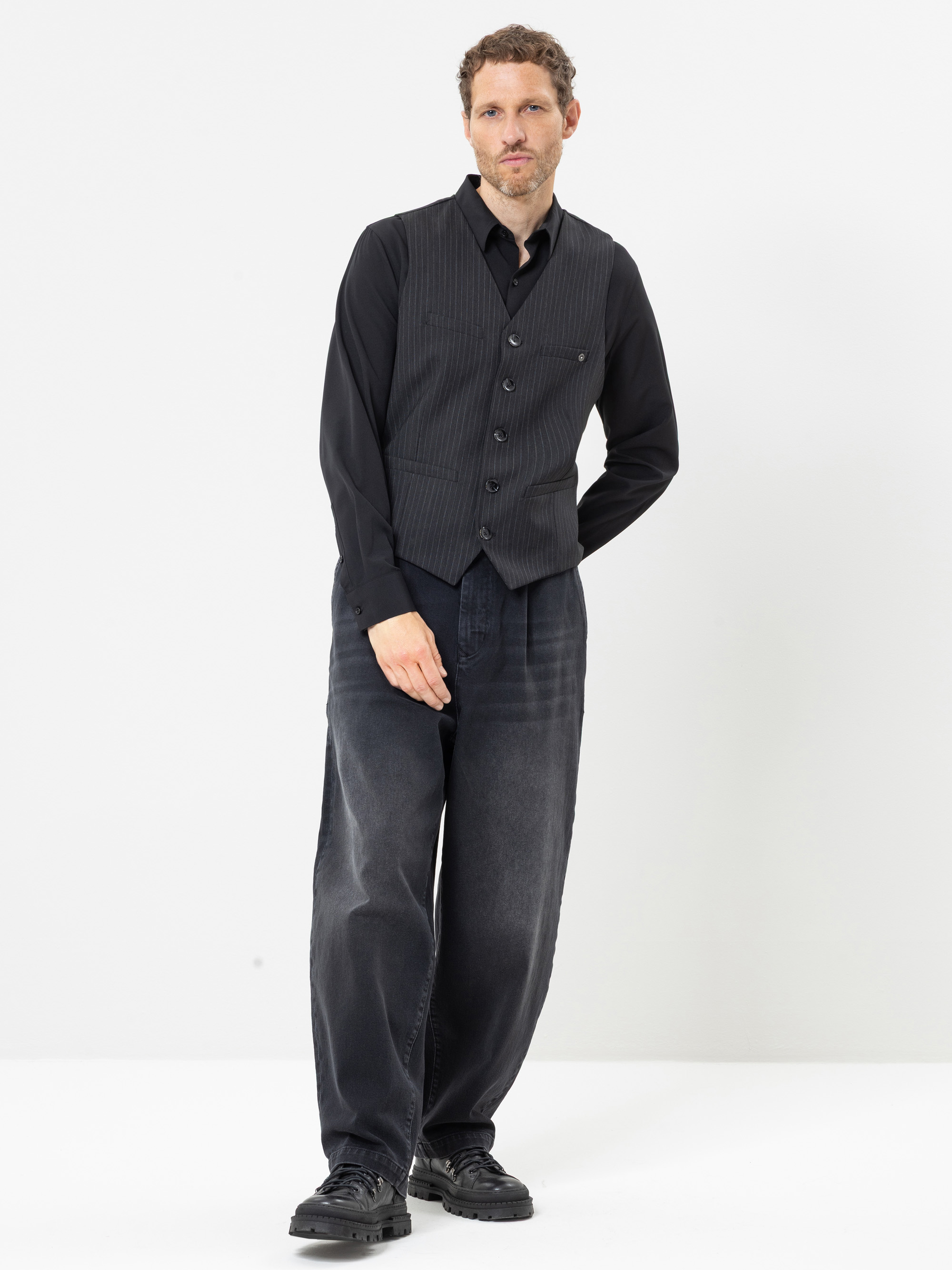 Ein Mann mit kurzen Locken trägt ein schwarzes Hemd mit Knöpfen, eine dunkelgraue Weste mit Nadelstreifen, eine weite dunkle Jeans und schwarze Schuhe; er steht vor einem schlichten weißen Hintergrund und hat eine Hand in der Hosentasche.