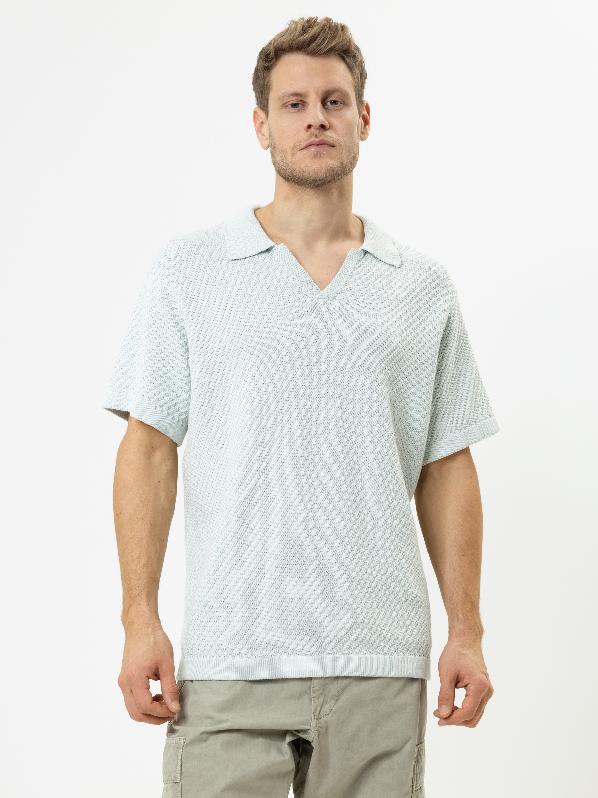 Ein Mann steht vor einem schlichten weißen Hintergrund und trägt ein hellblaues, strukturiertes, kurzärmeliges Hemd mit breitem Kragen und eine hellgraue Cargohose. Er blickt mit neutralem Blick geradeaus.