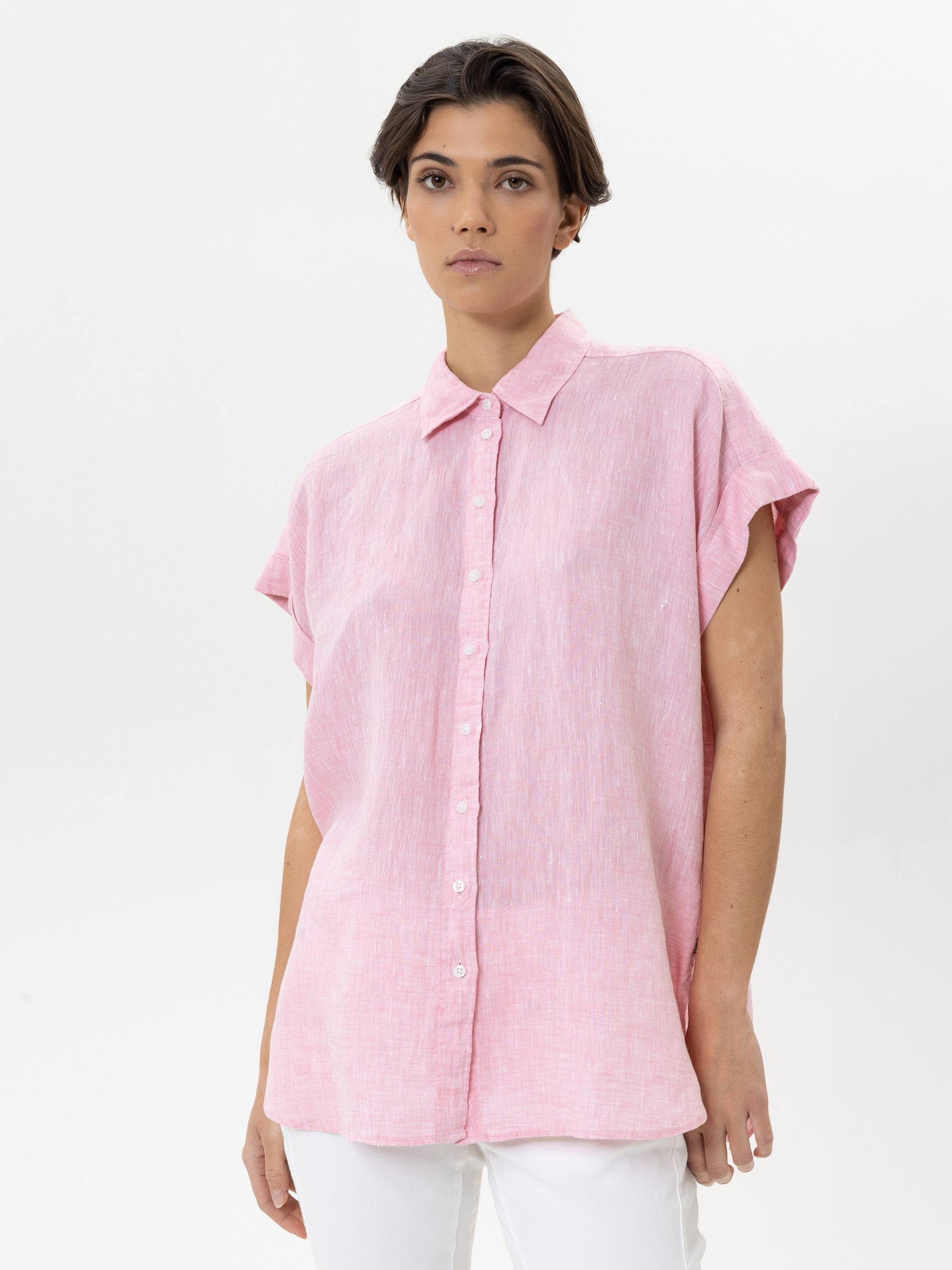 Eine Person mit kurzen braunen Haaren trägt ein locker sitzendes, kurzärmeliges rosa Button-up-Hemd und eine weiße Hose und steht vor einem schlichten weißen Hintergrund.