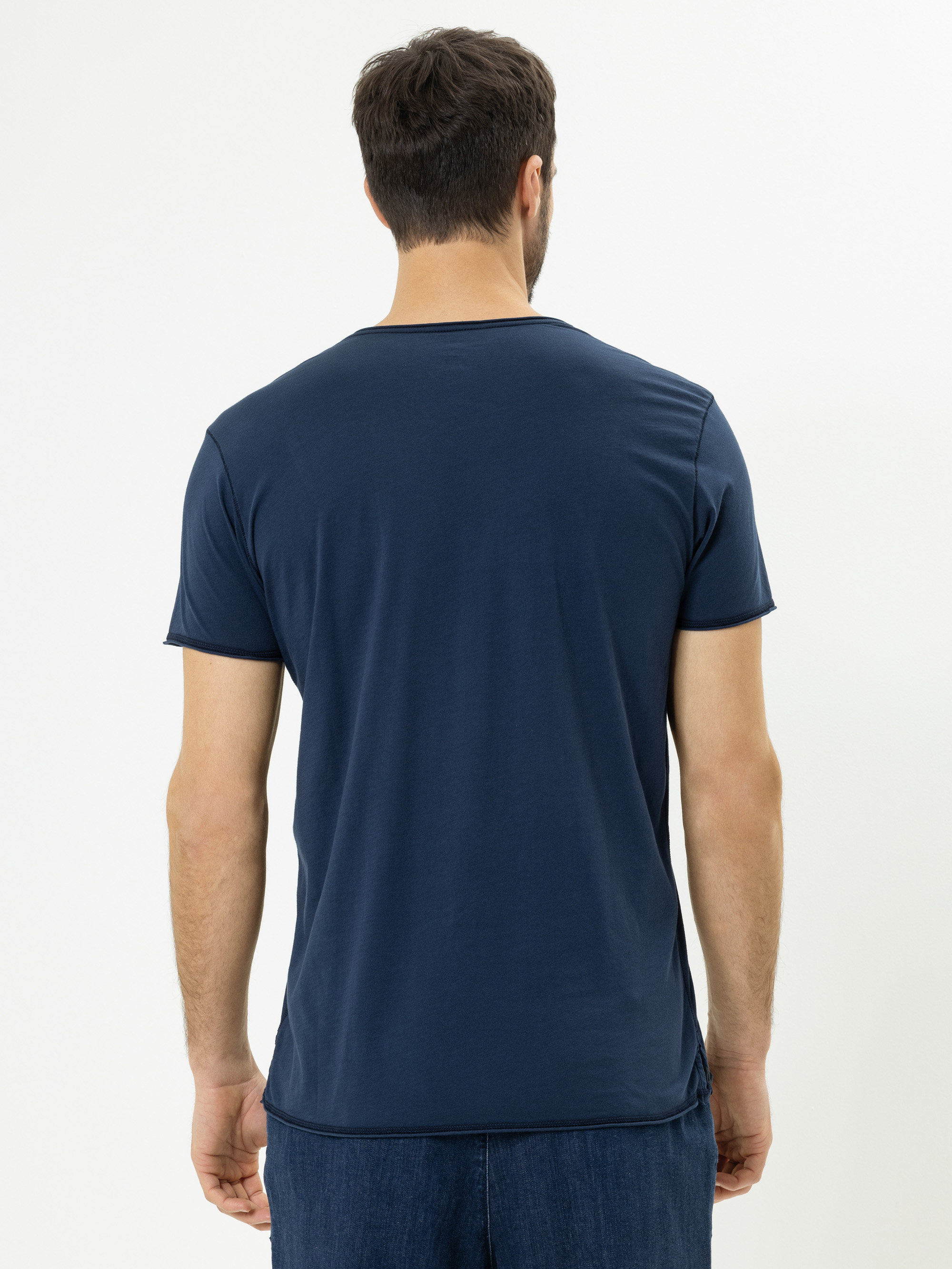 Ein Mann mit kurzen dunklen Haaren steht von der Kamera abgewandt und trägt ein schlichtes marineblaues T-Shirt und eine blaue Jeans vor einem weißen Hintergrund.