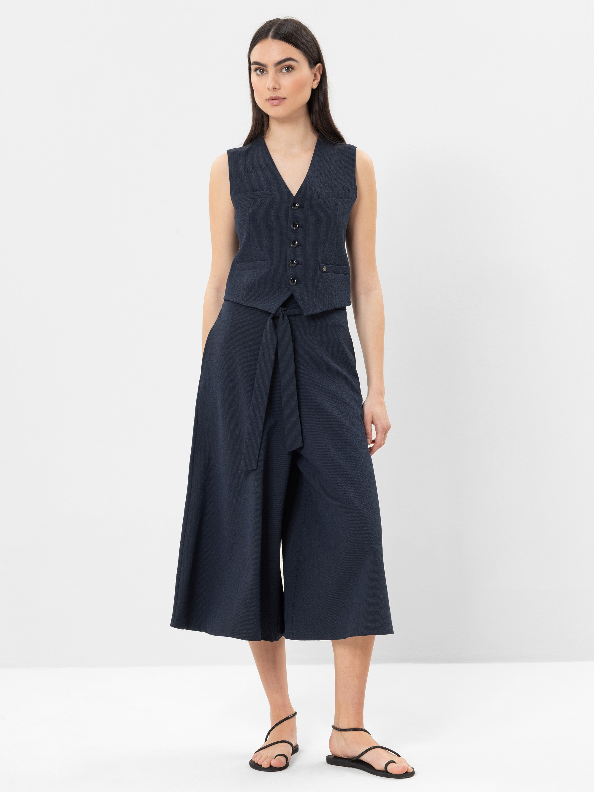 Eine Frau steht vor einem weißen Hintergrund und trägt eine ärmellose marineblaue Weste mit Knöpfen, eine weite marineblaue Culotte-Hose mit Bindegürtel und schwarze Sandalen. Sie hat glattes dunkles Haar und einen neutralen Gesichtsausdruck.