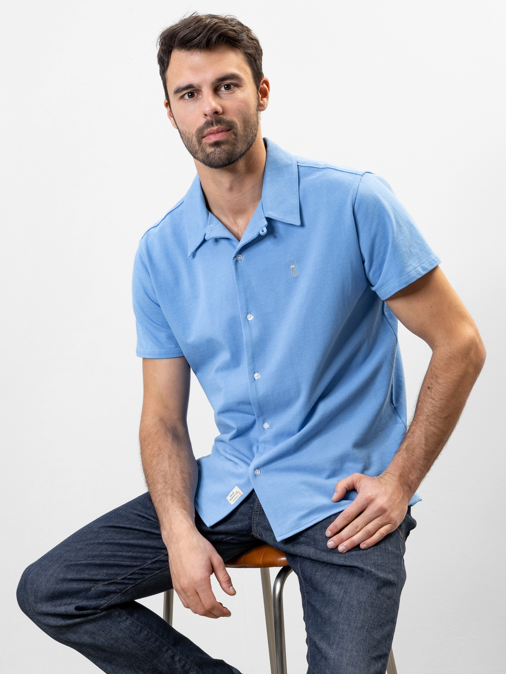 Ein Mann mit kurzen dunklen Haaren und Bart sitzt auf einem Hocker, trägt ein hellblaues, kurzärmeliges Button-up-Hemd und eine dunkle Jeans und blickt mit neutralem Blick in die Kamera vor einem schlichten weißen Hintergrund.