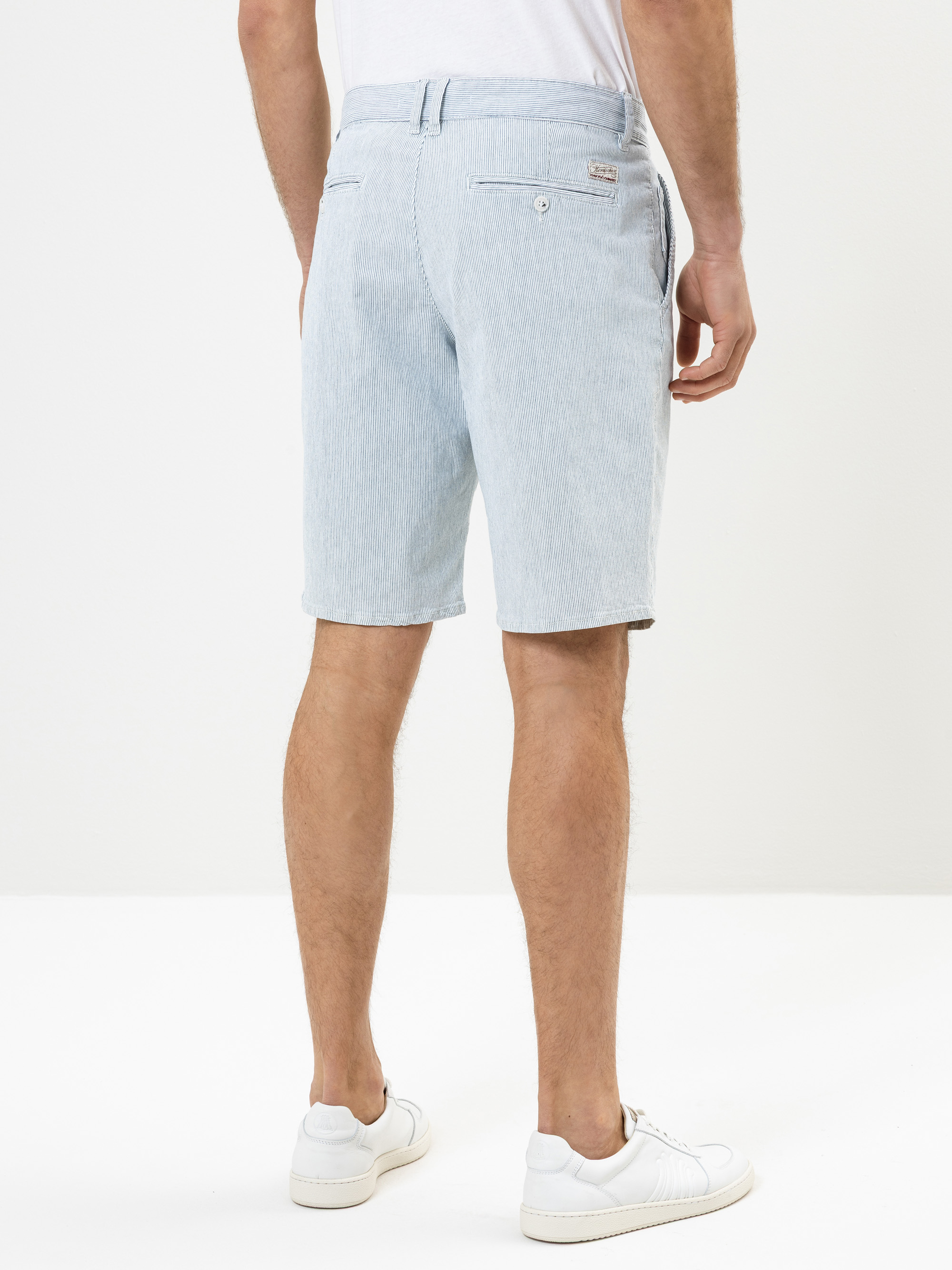 Ein Mann steht in hellblau gestreiften Shorts, einem weißen T-Shirt und weißen Turnschuhen vor einem schlichten weißen Hintergrund.