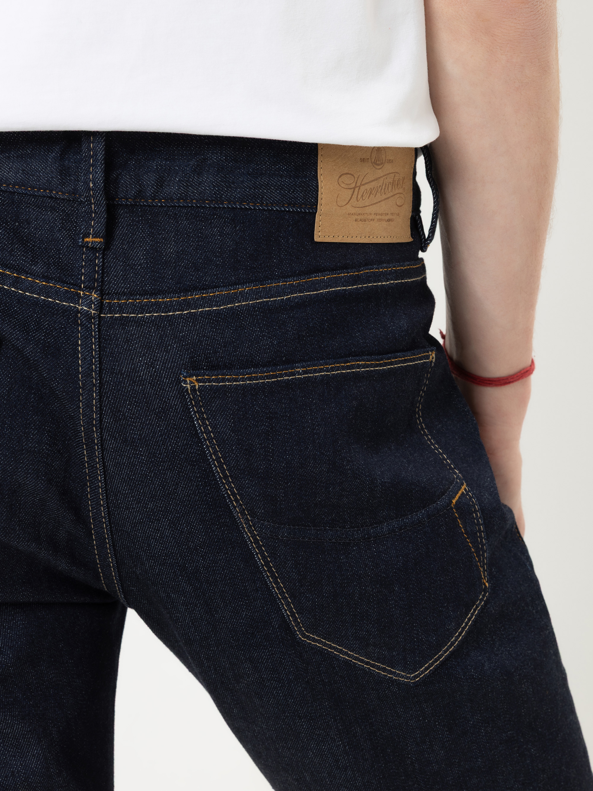 Eine Person, die dunkelblaue Jeans und ein weißes Hemd trägt, ist von hinten zu sehen. Die Jeans hat gelbe Nähte und einen braunen Lederaufnäher am Bund, und die Person trägt ein rotes Schnurarmband am Handgelenk.