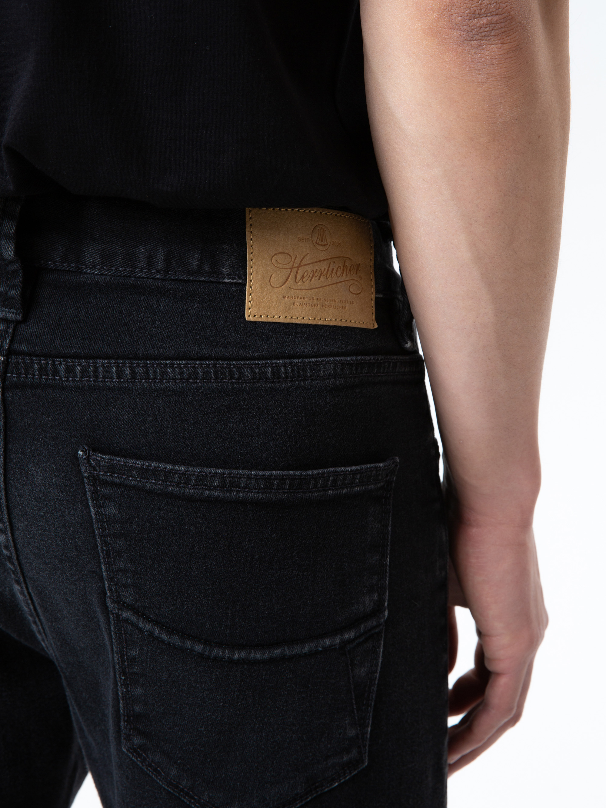 Eine Person, die schwarze Jeans und ein schwarzes Hemd trägt, wird von hinten gezeigt. Die Jeans hat am Bund einen braunen Lederaufnäher mit der Aufschrift Heppitcher.