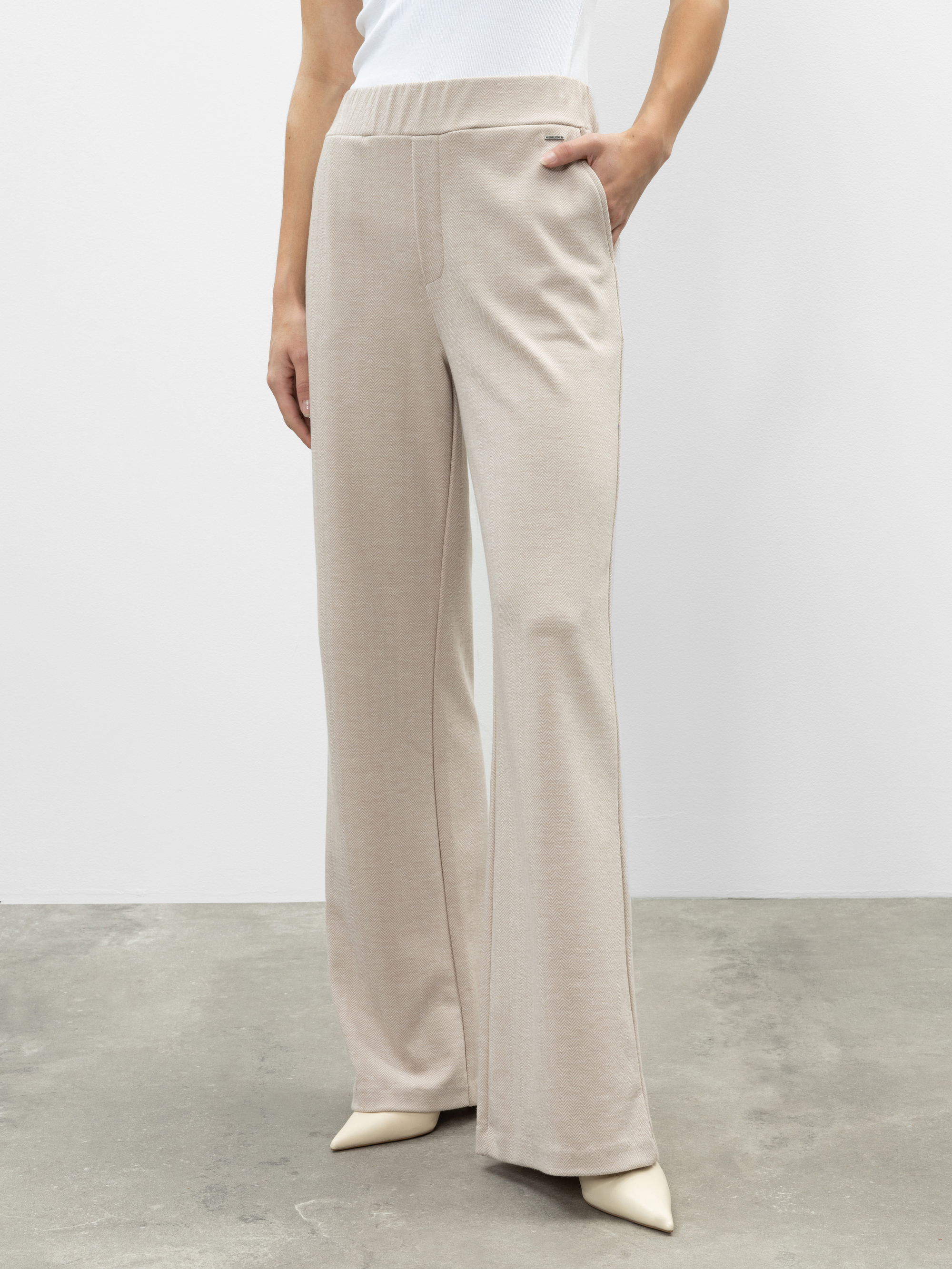 Eine Person, die eine hellbeige, weit geschnittene Hose mit elastischem Bund und ein weißes Oberteil trägt; ihre rechte Hand steckt in der Tasche und sie steht auf einem grauen Betonboden.