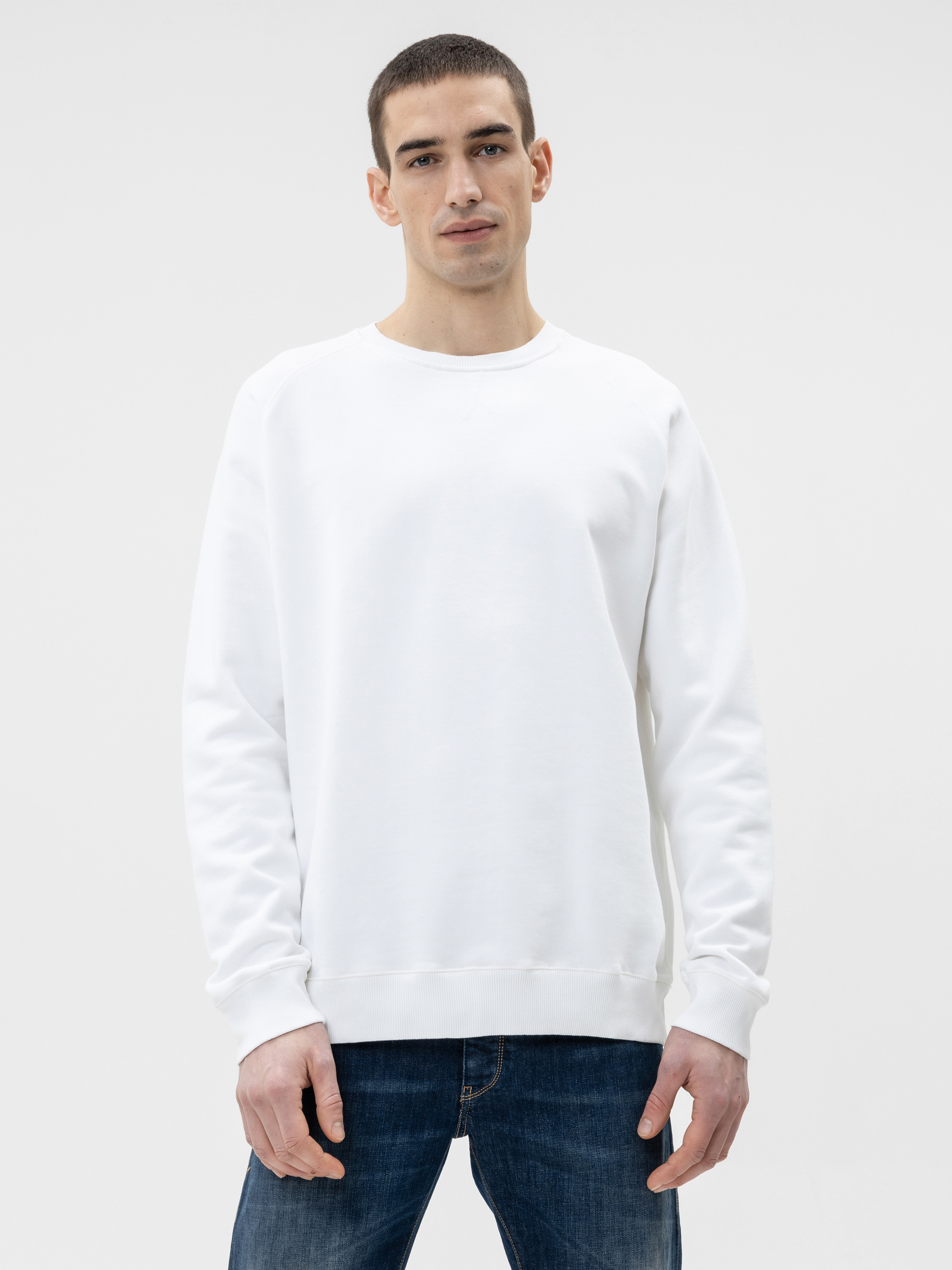 Ein Mann mit kurzen dunklen Haaren, der ein einfaches weißes Sweatshirt und blaue Jeans trägt, steht vor einem einfachen weißen Hintergrund und blickt mit neutralem Blick in die Kamera.