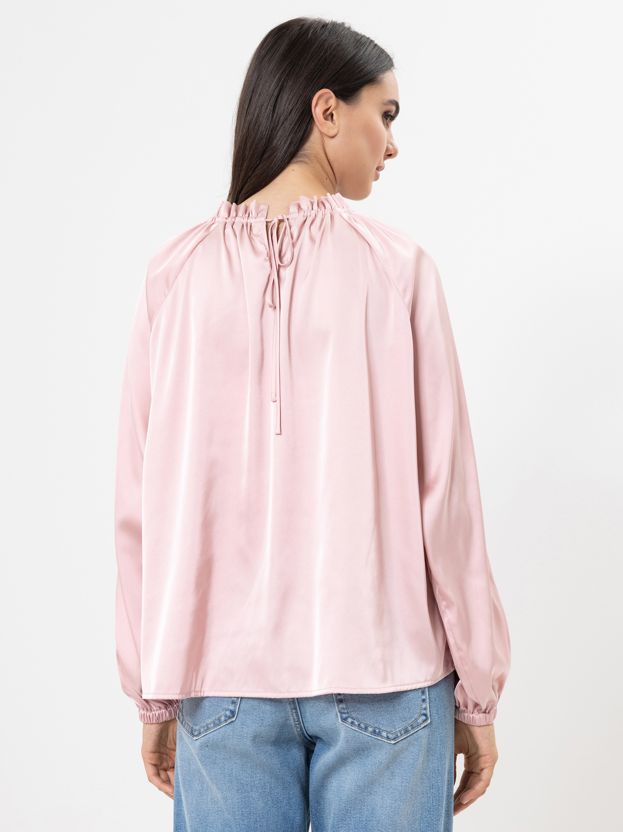 Eine Frau mit langen dunklen Haaren trägt eine lockere, hellrosa Satinbluse mit Rüschenausschnitt und Bindeband im Rücken, dazu eine hellblaue Jeans. Sie steht und schaut von der Kamera weg.