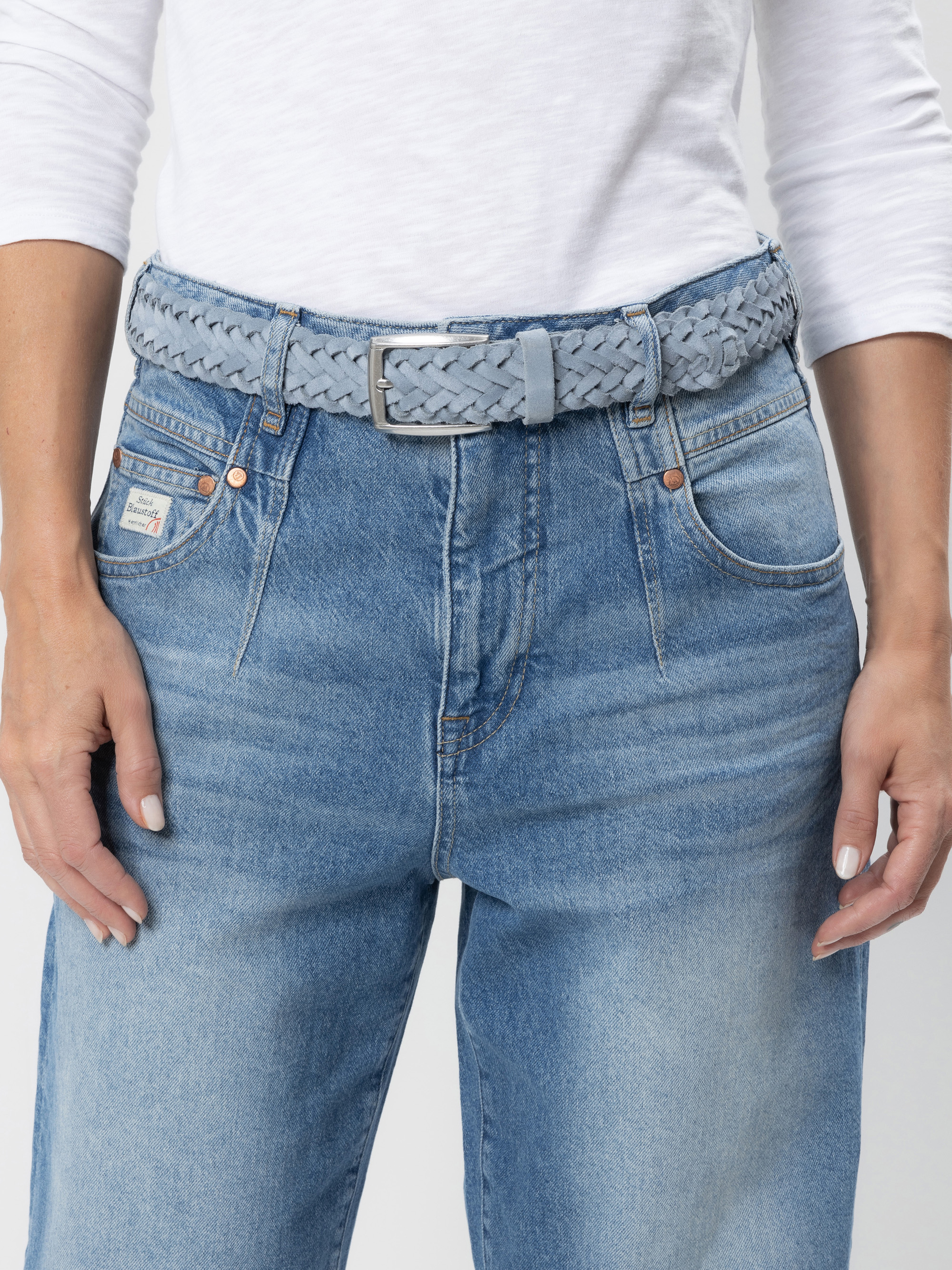 Eine Person, die ein weißes, langärmeliges Hemd trägt, das in eine hochgekrempelte blaue Jeans mit Vordertaschen und einem geflochtenen hellblauen Gürtel gesteckt ist. Die Hände liegen entspannt an der Seite.