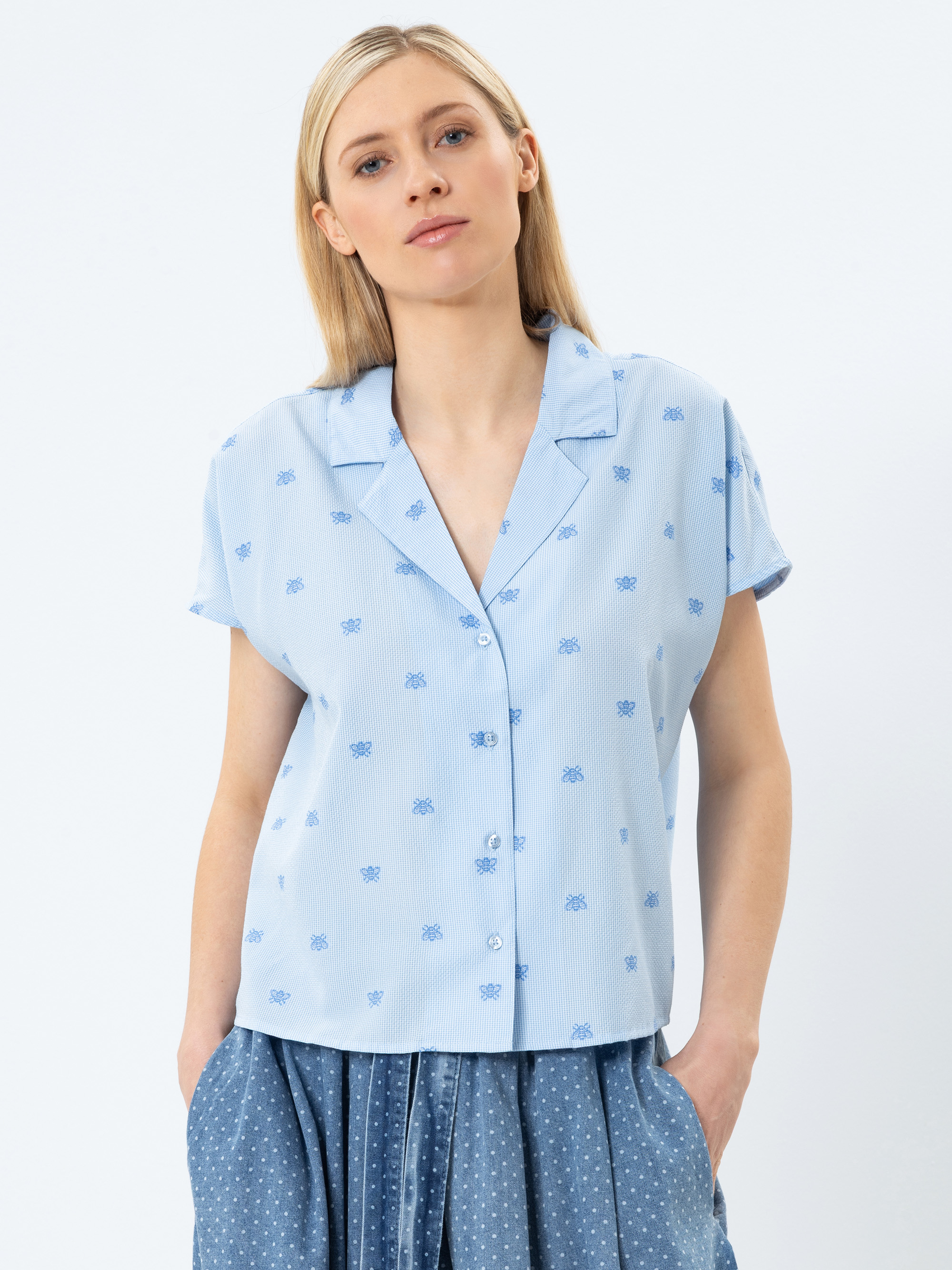 Eine Frau mit glattem blondem Haar trägt ein kurzärmeliges, hellblaues Button-up-Hemd mit einem dezenten blauen Aufdruck und einem Kragen, gepaart mit einem blauen gepunkteten Rock. Sie steht mit den Händen in ihren Rocktaschen vor einem weißen Hinter