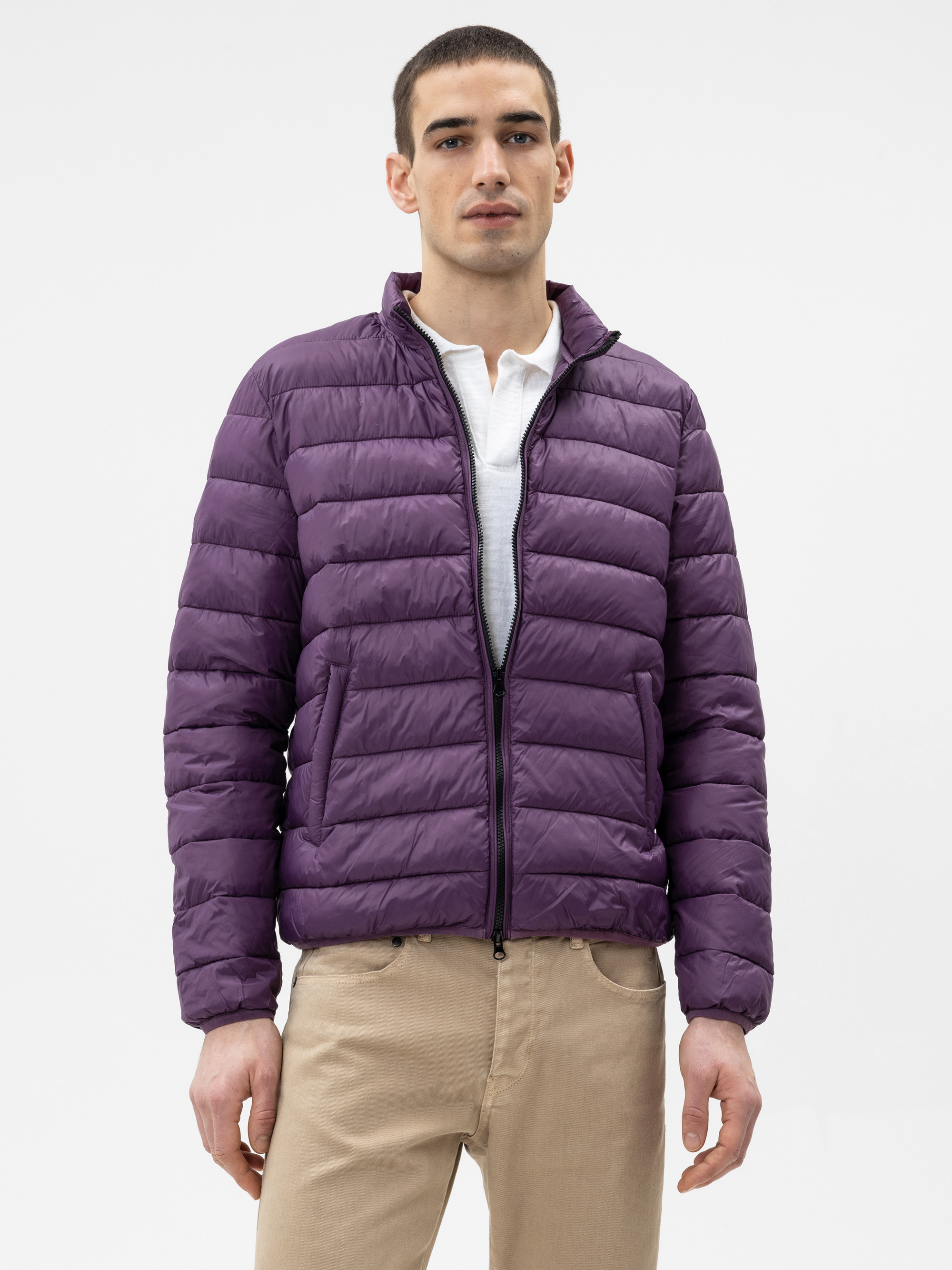 Ein Mann steht mit dem Gesicht nach vorne, trägt eine lila Pufferjacke über einem weißen Hemd mit Kragen und eine beigefarbene Hose vor einem schlichten hellen Hintergrund.