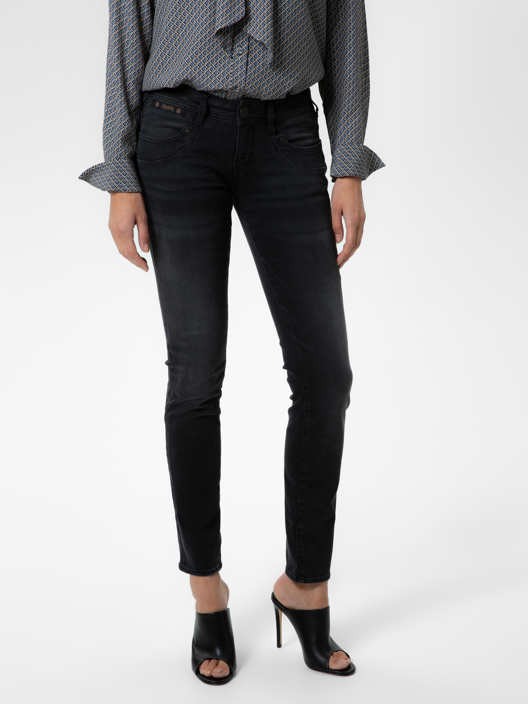 Eine Person, die dunkle Röhrenjeans, eine gemusterte langärmelige Bluse und schwarze Pantoletten mit offenen Zehen trägt, steht vor einem schlichten weißen Hintergrund. Nur der Körper von den Schultern abwärts ist sichtbar.