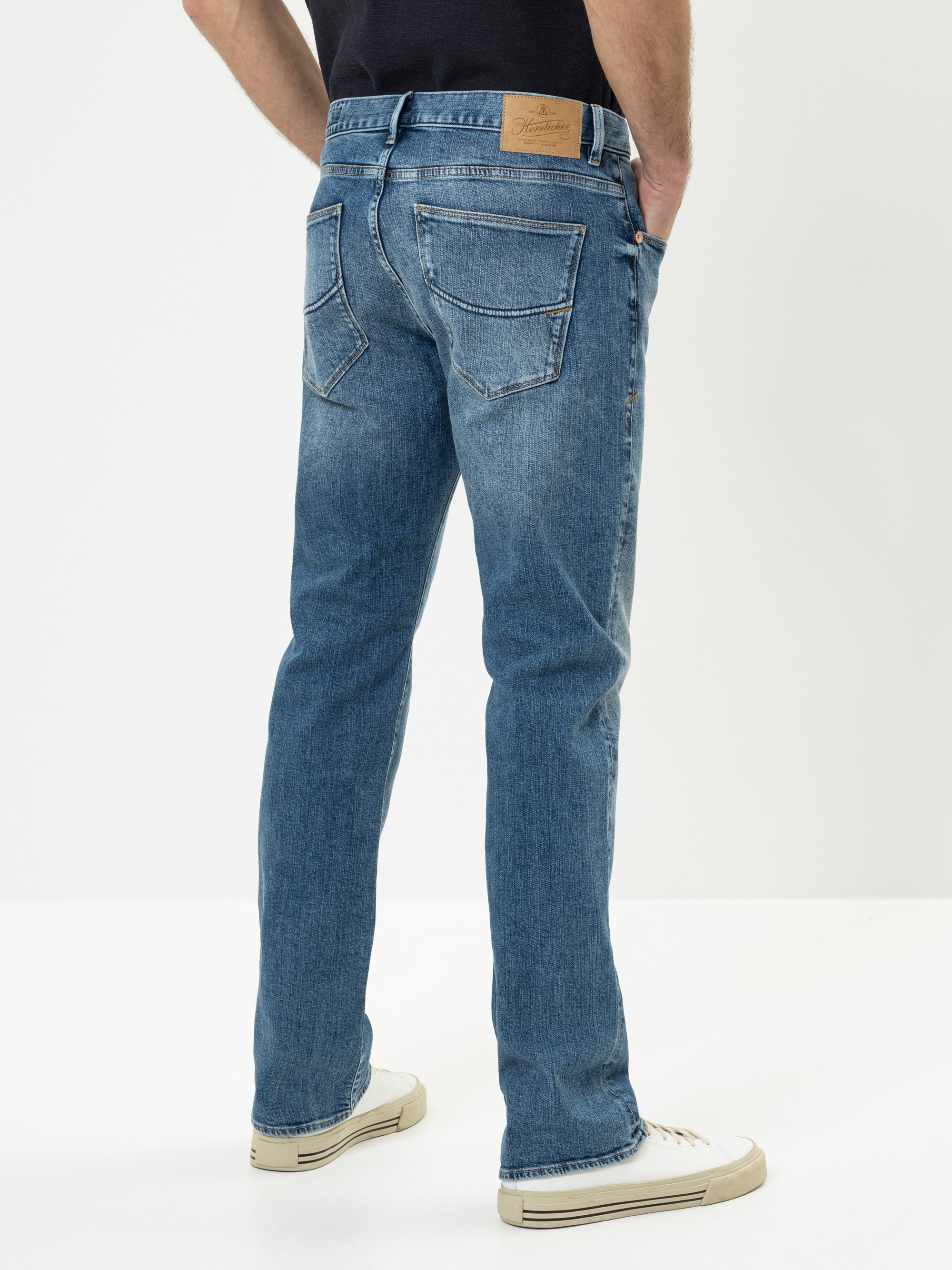 Eine Person steht mit dem Gesicht zur Wand und trägt eine blaue Jeans mit sichtbaren Gesäßtaschen und einem hellbraunen Lederaufnäher am Bund, weiße Turnschuhe und ein schwarzes Hemd. Der Hintergrund ist einfarbig und hell.