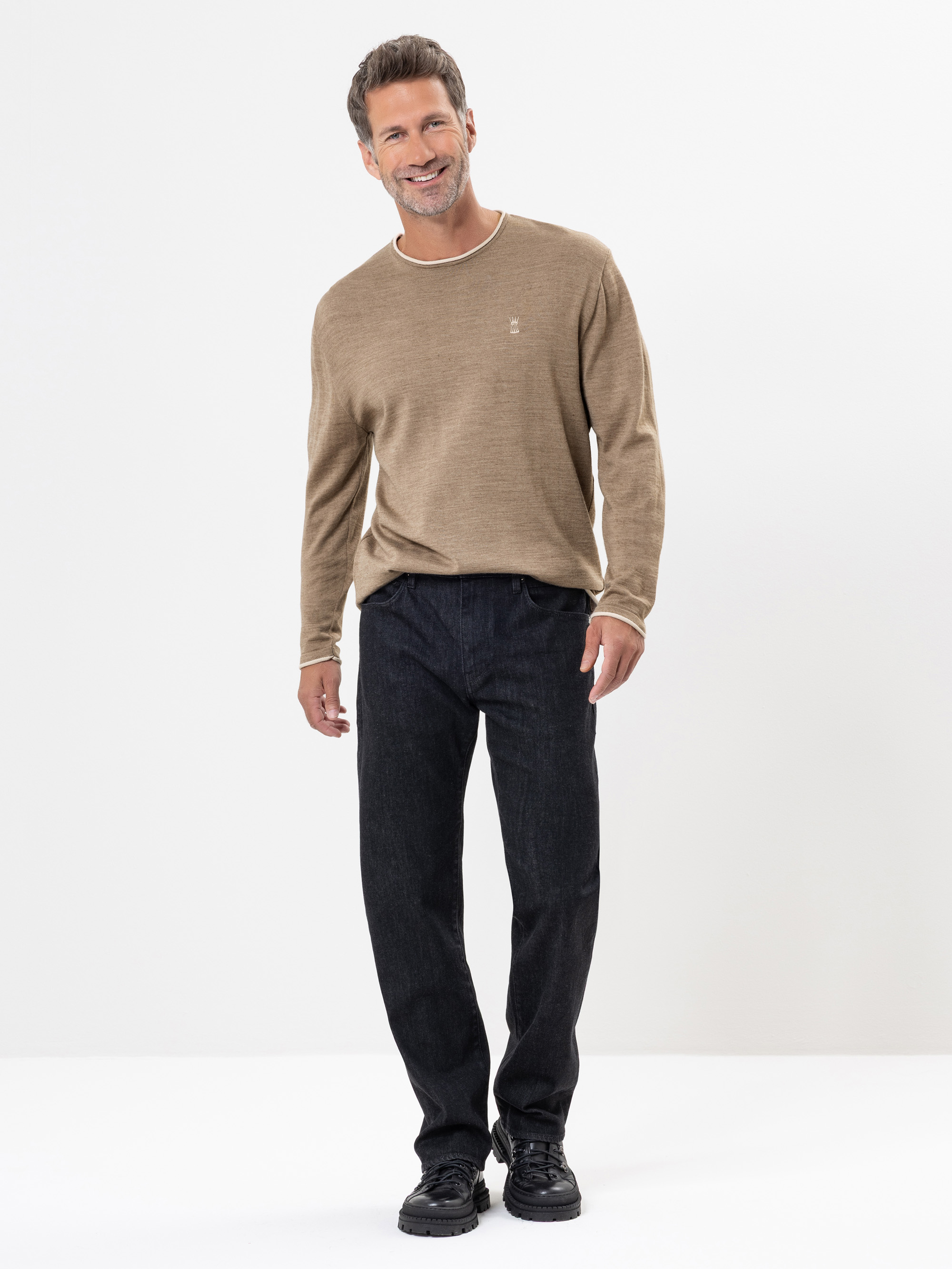 Ein Mann mit kurzen braunen Haaren und einem Bart lächelt im Stehen. Er trägt einen hellbraunen langärmeligen Pullover, dunkle Jeans und schwarze Schuhe vor einem schlichten weißen Hintergrund.