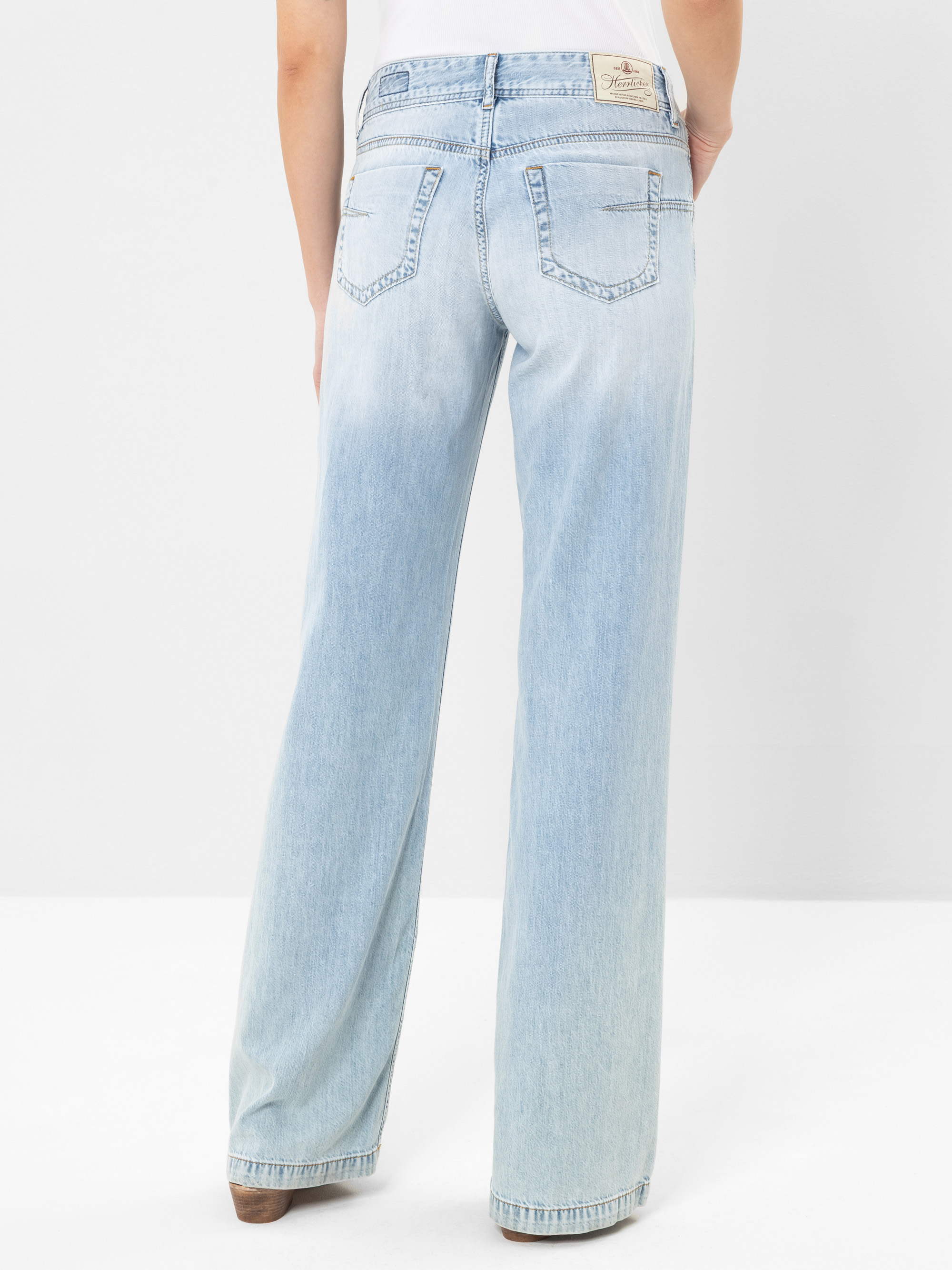 Eine Person, die eine helle, weit geschnittene Jeans und braune Schuhe trägt, steht mit dem Gesicht nach hinten und zeigt die Gesäßtaschen und ein Etikett am Bund vor einem schlichten weißen Hintergrund.