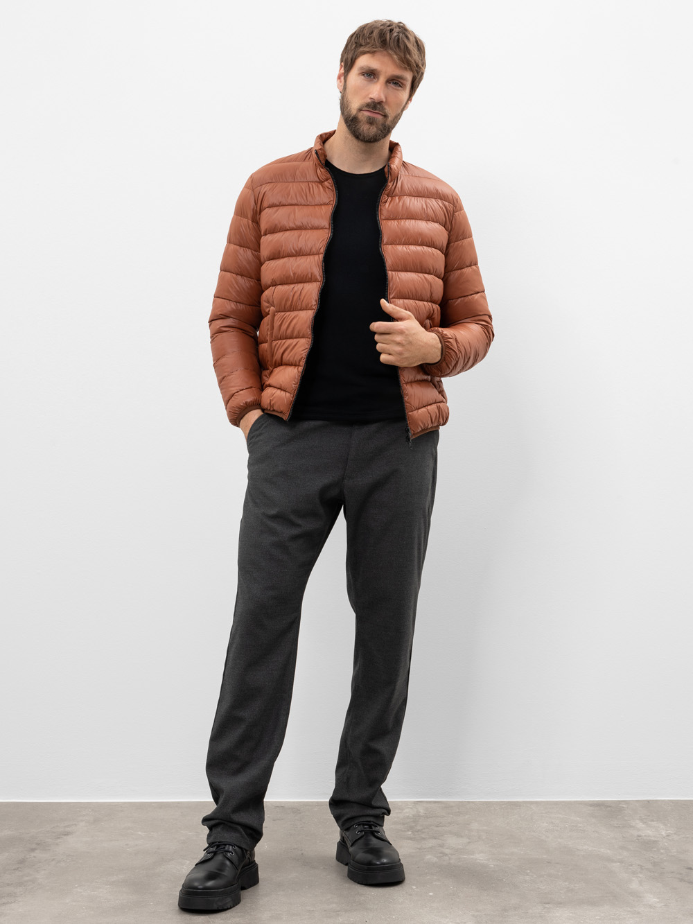Ein Mann mit kurzen braunen Haaren und einem Bart trägt eine braune Pufferjacke, ein schwarzes Hemd, eine graue Hose und schwarze Schuhe und steht vor einem schlichten weißen Hintergrund auf einem hellgrauen Boden.