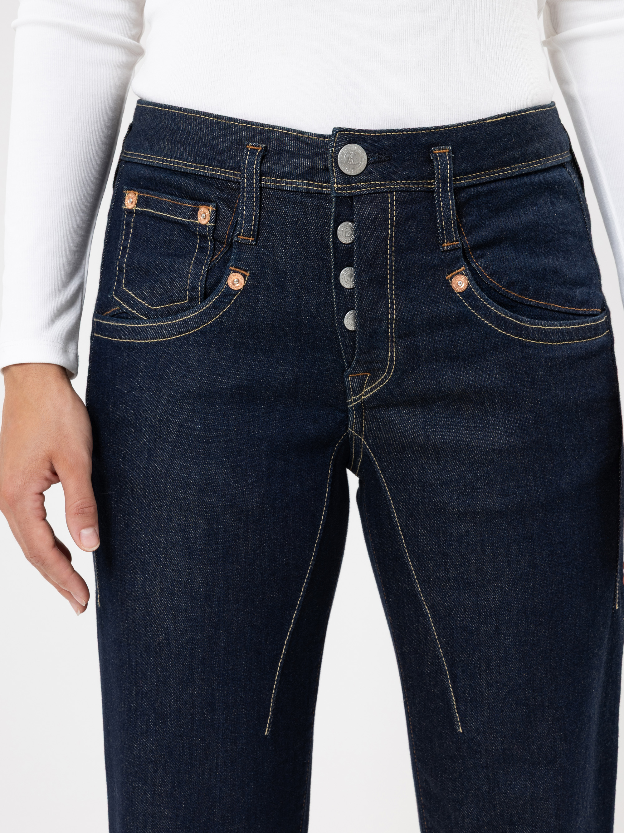 Eine Person trägt eine dunkelblaue Jeans mit freiliegenden silbernen Knöpfen und ein weißes langärmeliges Oberteil, das von der Taille bis zur Mitte des Oberschenkels gezeigt wird.