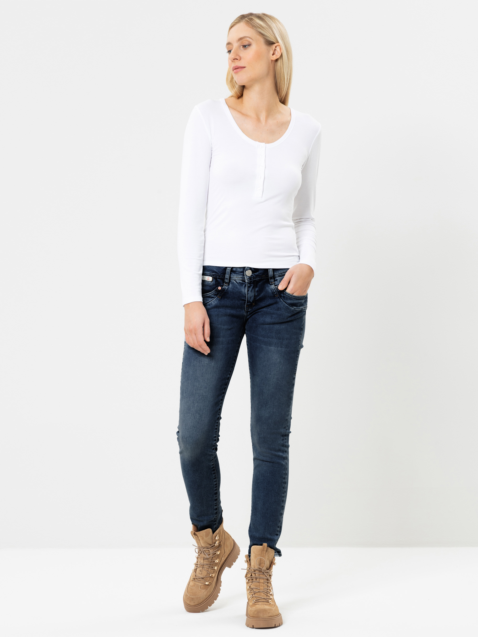 Eine Frau steht vor einem schlichten weißen Hintergrund und trägt ein weißes Langarmshirt, dunkelblaue Jeans und hellbraune Schnürstiefel. Sie hat blondes Haar und steckt ihre linke Hand in die Jeanstasche.