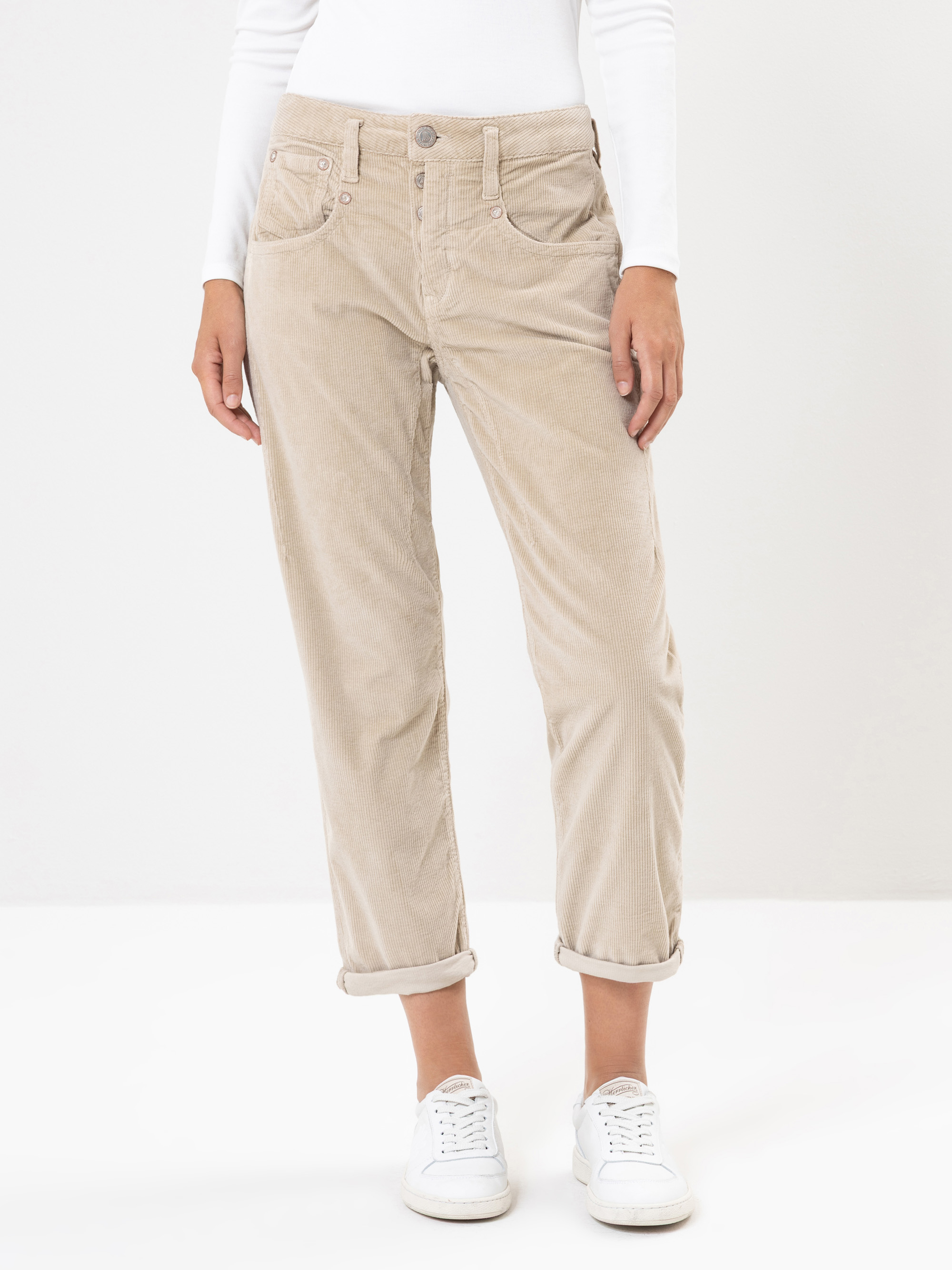 Eine Person, die eine hellbeige Cordhose mit aufgerollten Manschetten, ein weißes langärmeliges Oberteil und weiße Turnschuhe trägt, steht vor einem schlichten weißen Hintergrund.